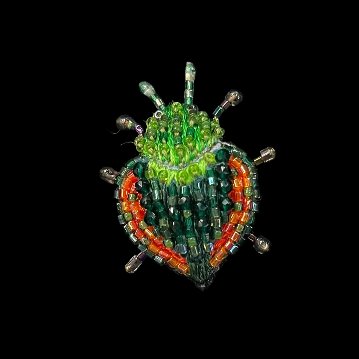 A handcrafted seed bead beetle brooch featuring a green head and body with orange-red accents along the sides, set against a black background.