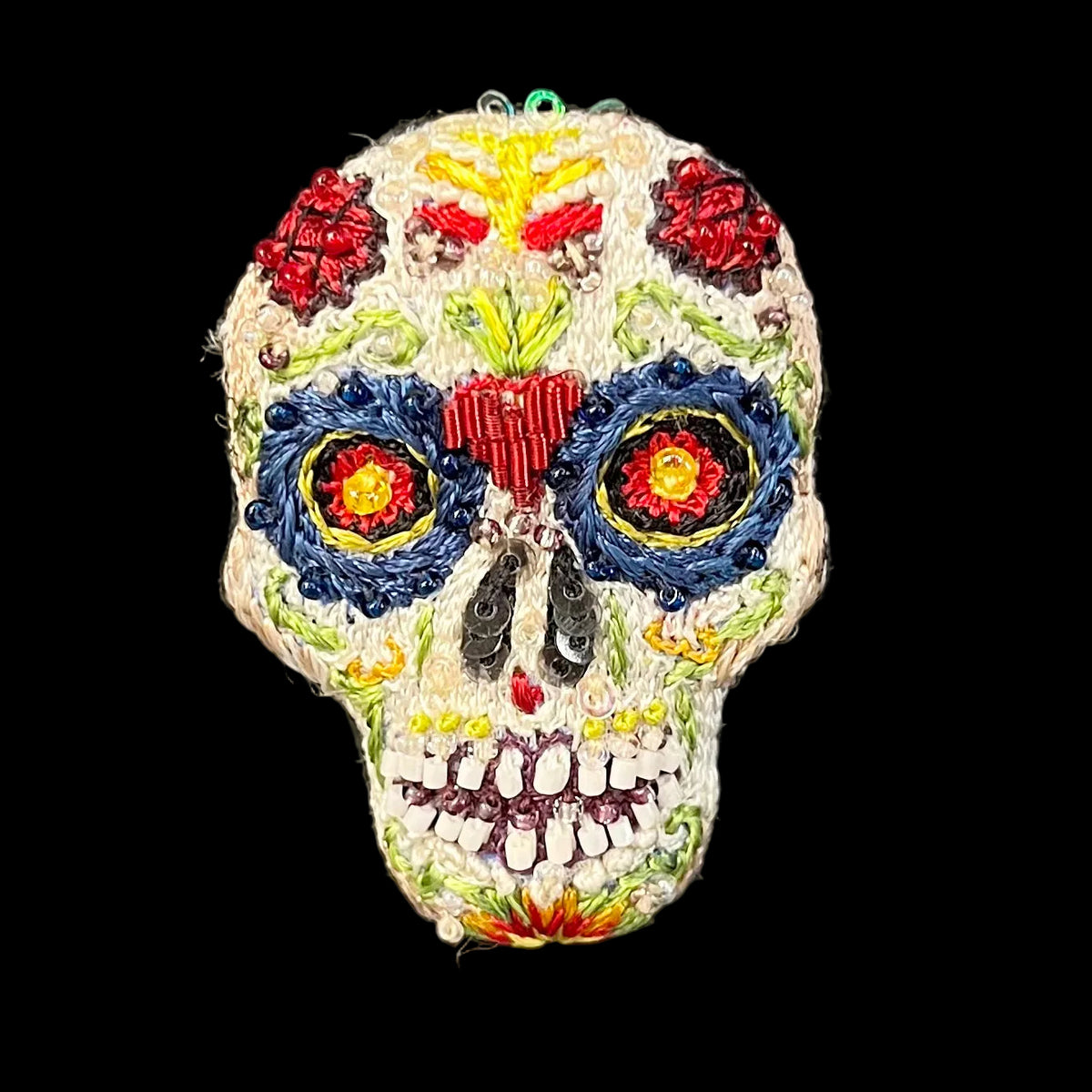 An embroidered sugar skull ornament featuring intricate floral and geometric patterns in red, blue, yellow, green, and black on a white base, set against a solid black background.