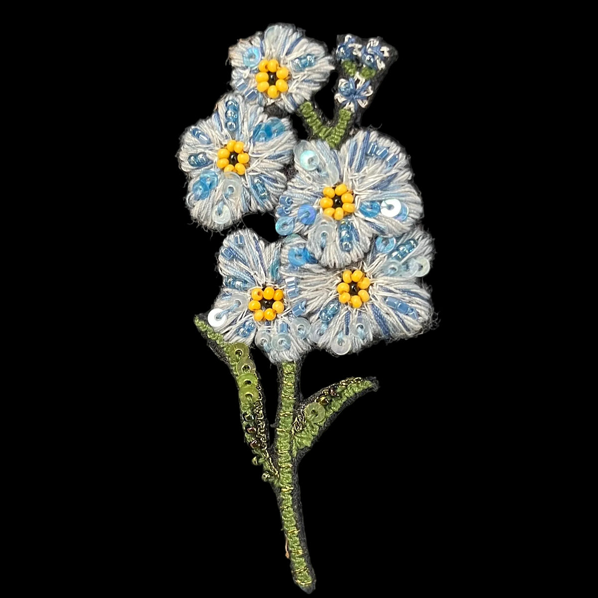 A handmade floral brooch featuring light blue and white paper beads with yellow centers and green stems, displayed on a black velvet surface.