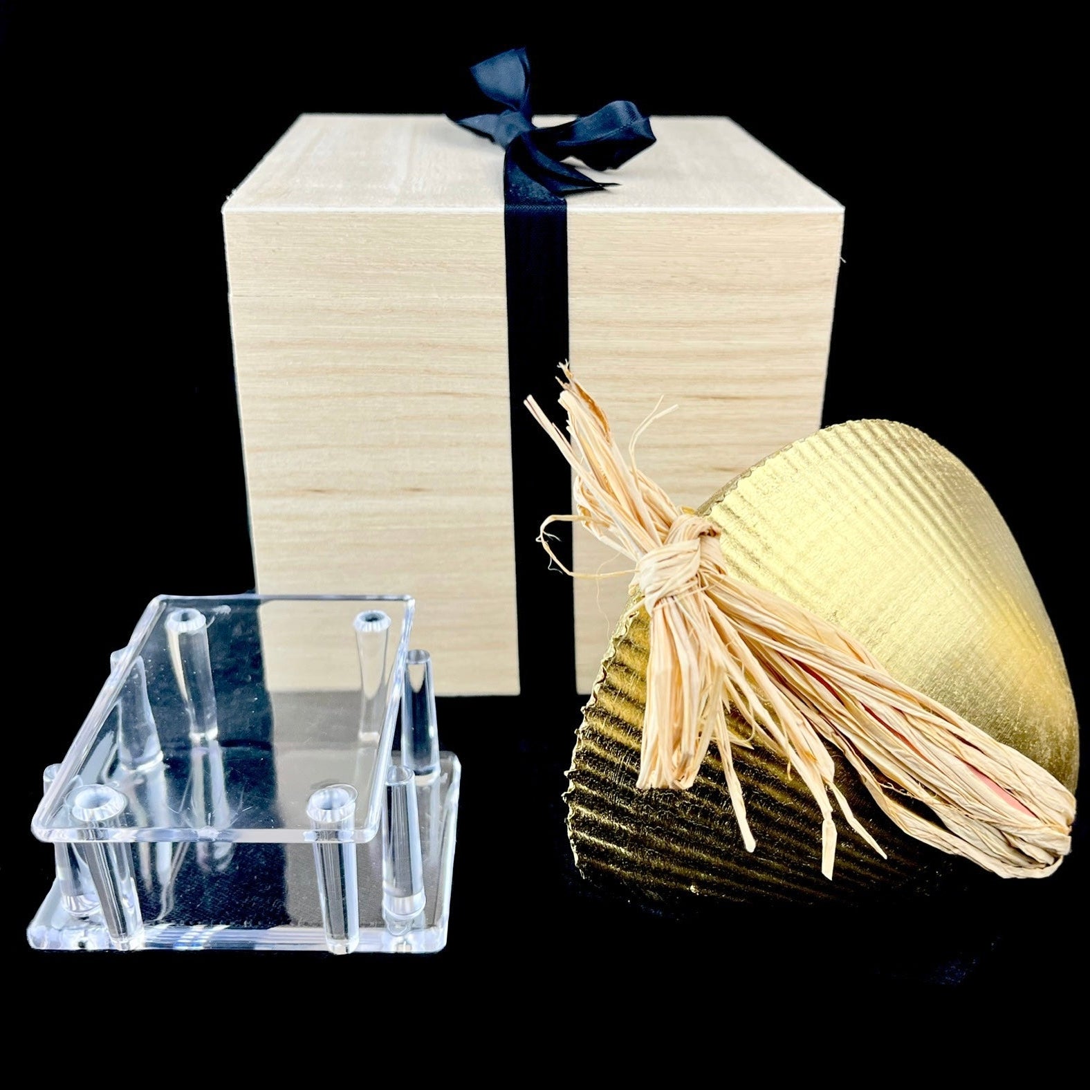 Decorative items including a gold ornament, clear stand, and gift box with a black ribbon on a black background.