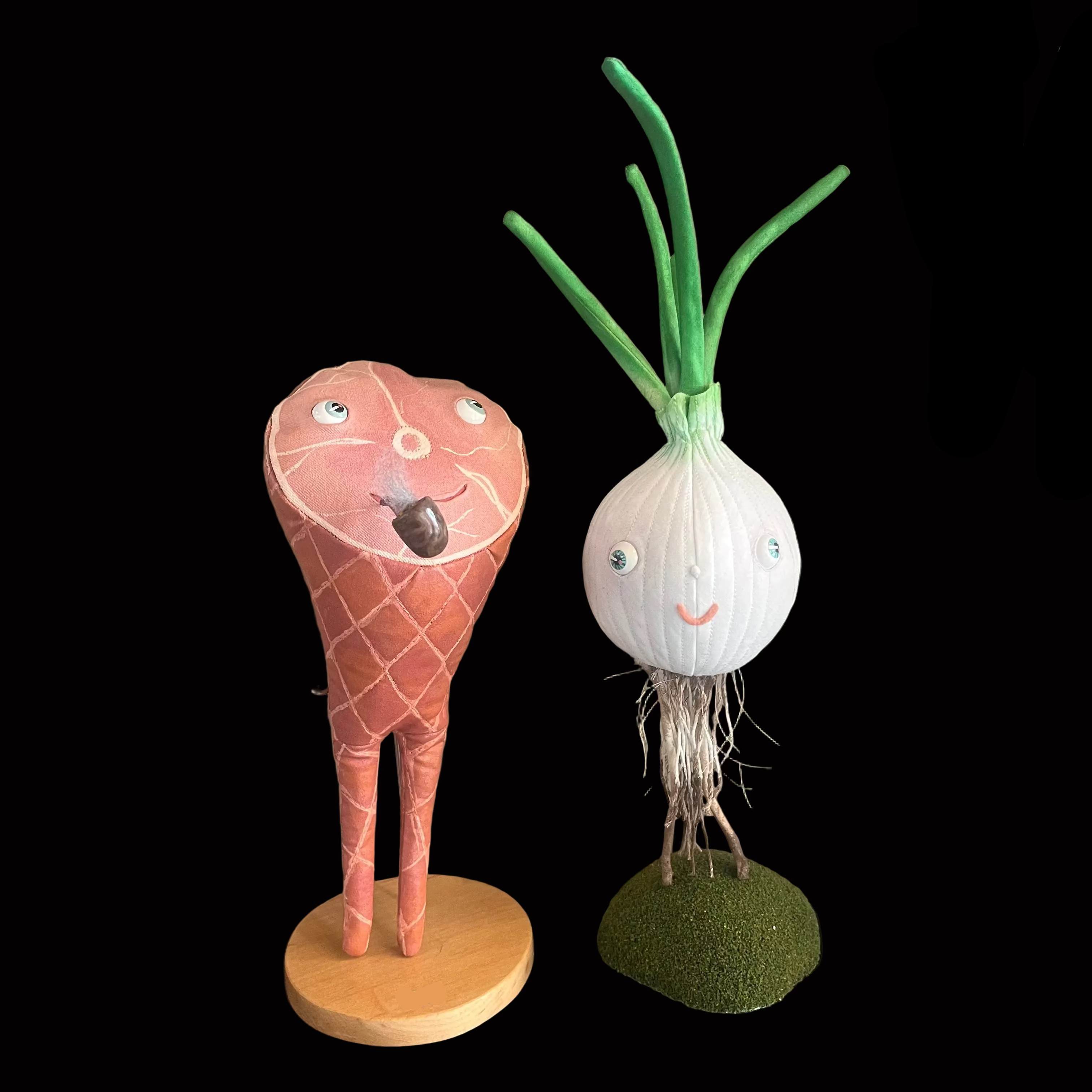 Whimsical smoked ham-shaped decorative figure standing on wooden base against black background with a smoking pipe and smiling face next to a white onion figure with green foliage and green turf stand.