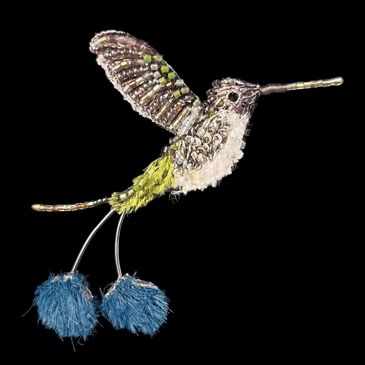 A handmade beaded and wrapped hummingbird brooch with blue pom-pom accents, displayed on a black velvet surface inside a blue box.
