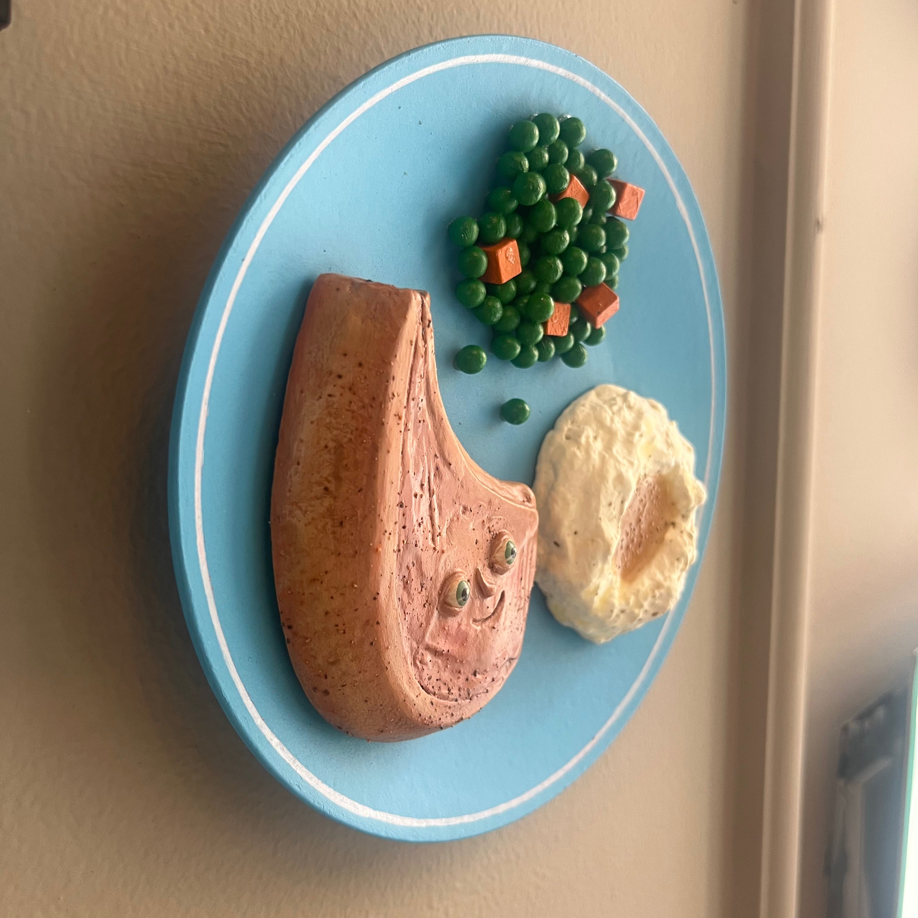 A wooden wall plaque shaped like a blue plate holding a cooked pork chop, mashed potatoes, and peas with small carrot cubes, on black background.