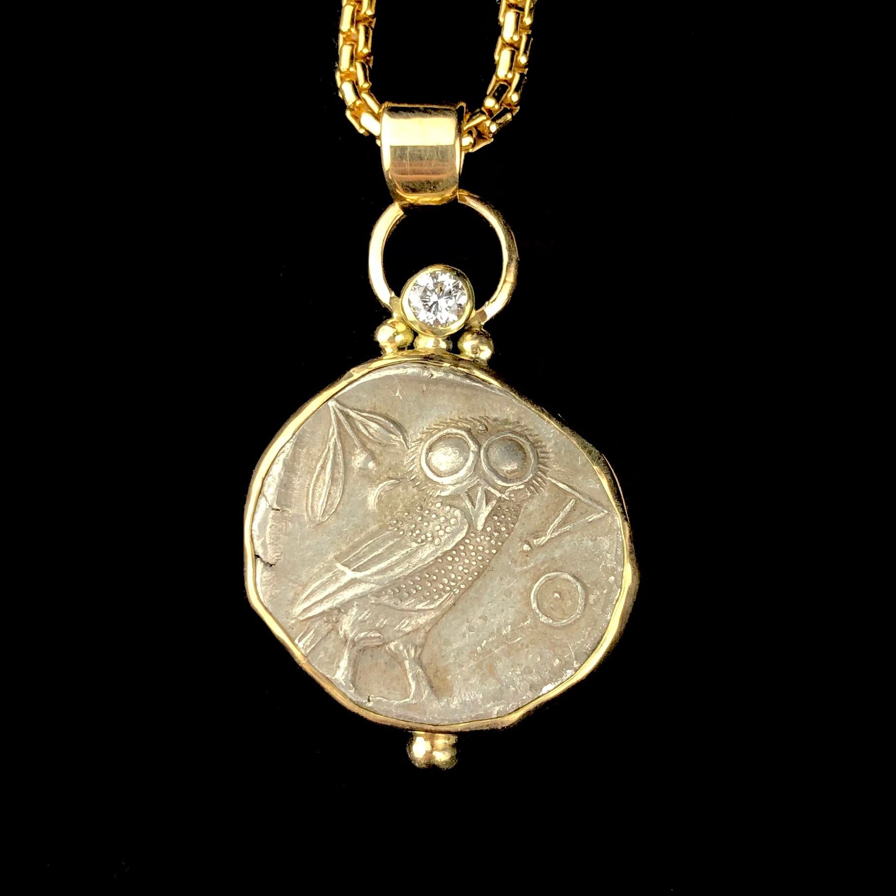An ancient greek silver coin with the relief of an owl set in gold with a large white diamond set at the top