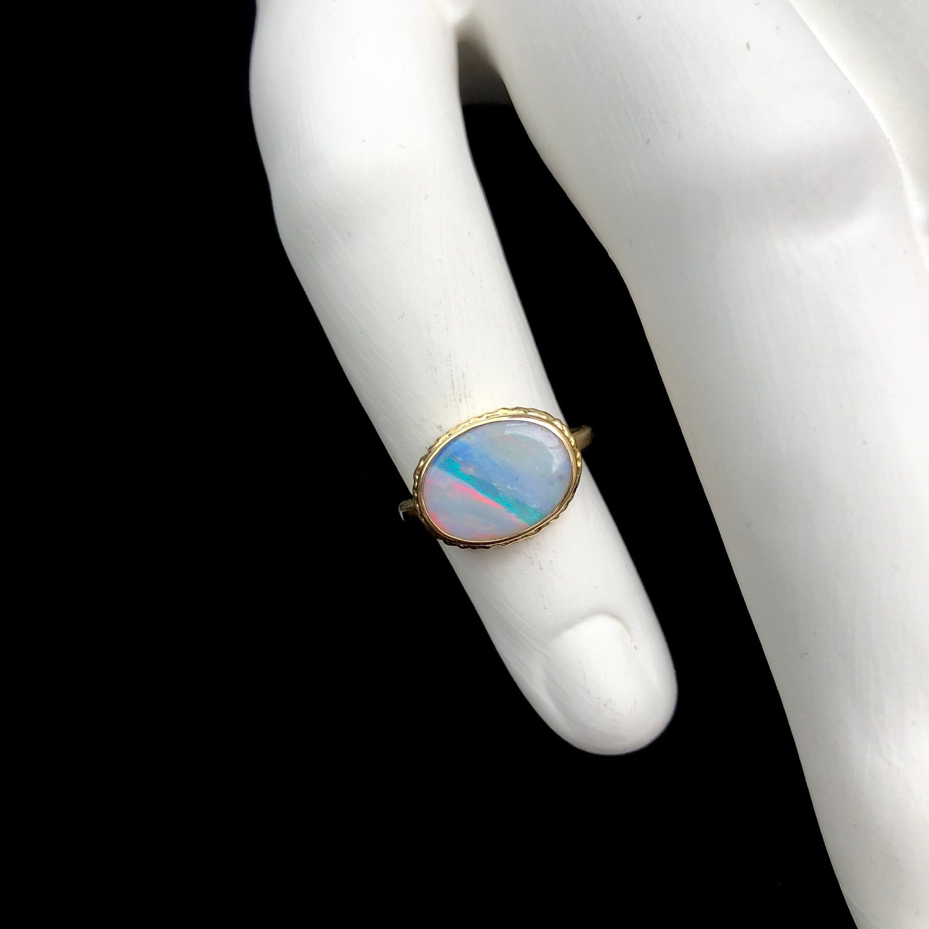 Top view of oval shaped white iridescent opal with green/blue stripe set in gold shown on white ceramic finger