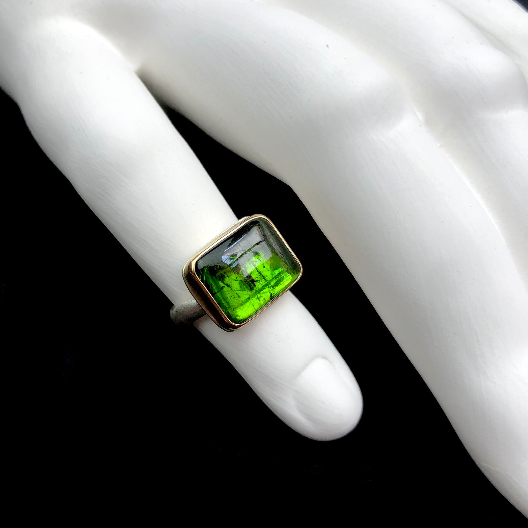 Green Tourmaline Ring with darker inclusions visible within the polished smooth stone shown on white ceramic finger