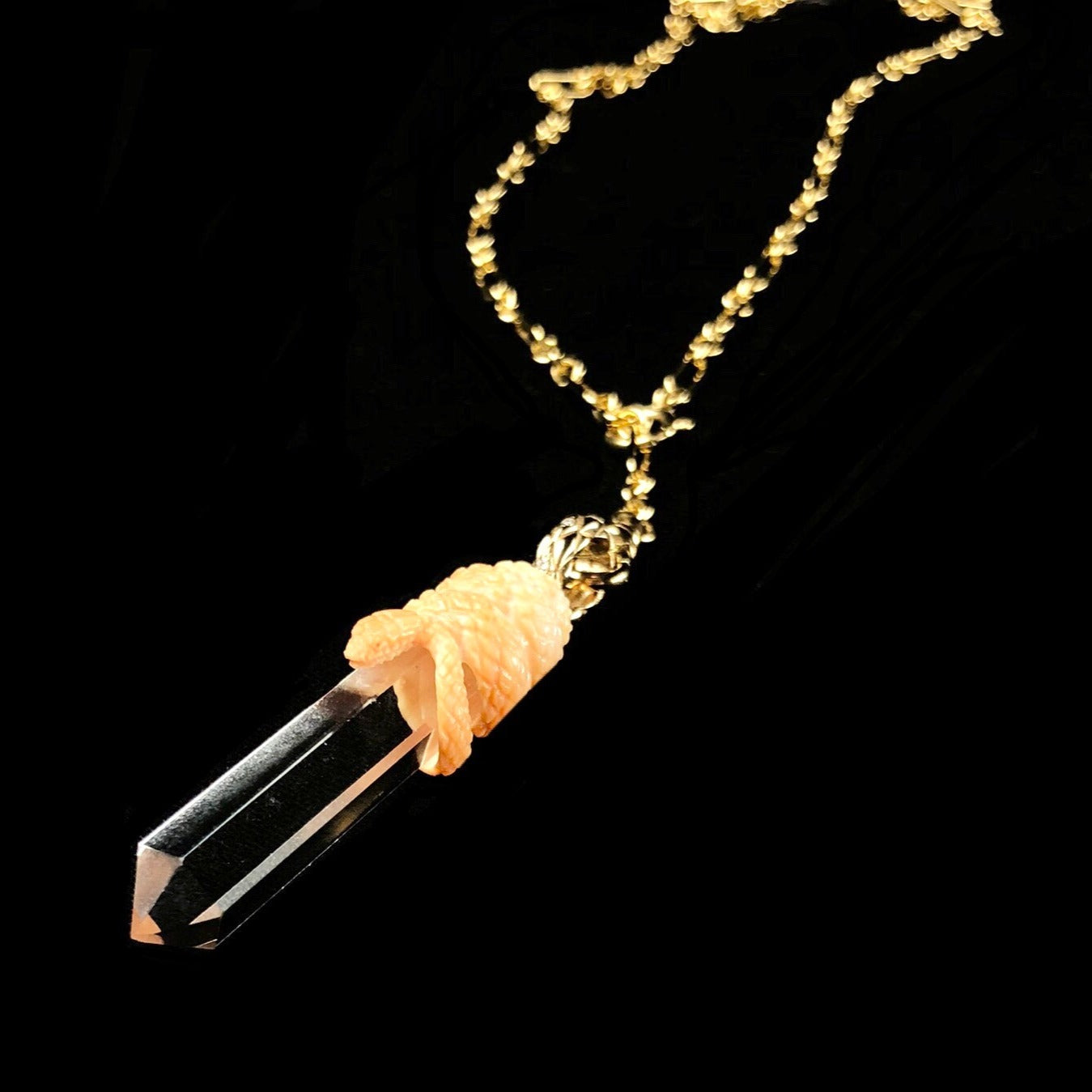 Clear crystal with light orange snake coiled about its top with gold bail with gold chain in background