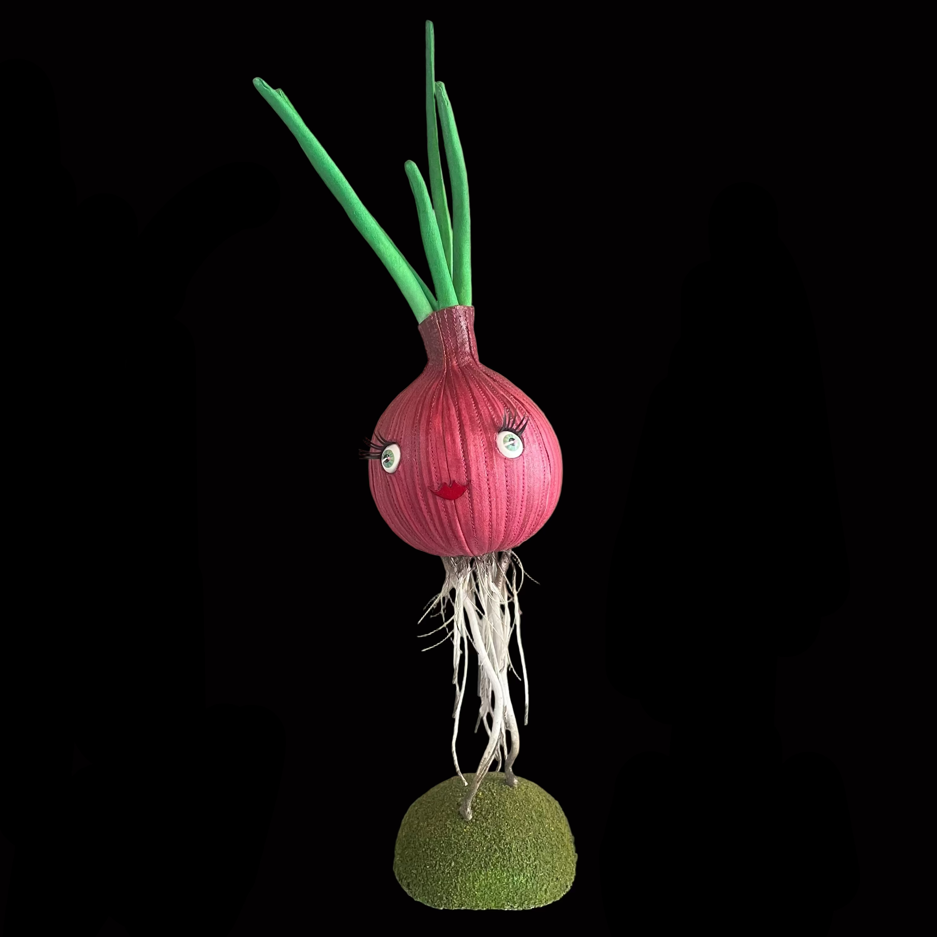 Whimsical vegetable-shaped decorative figure standing on mossy bases against black background resembling a red onion with green leaves and a smiling face.