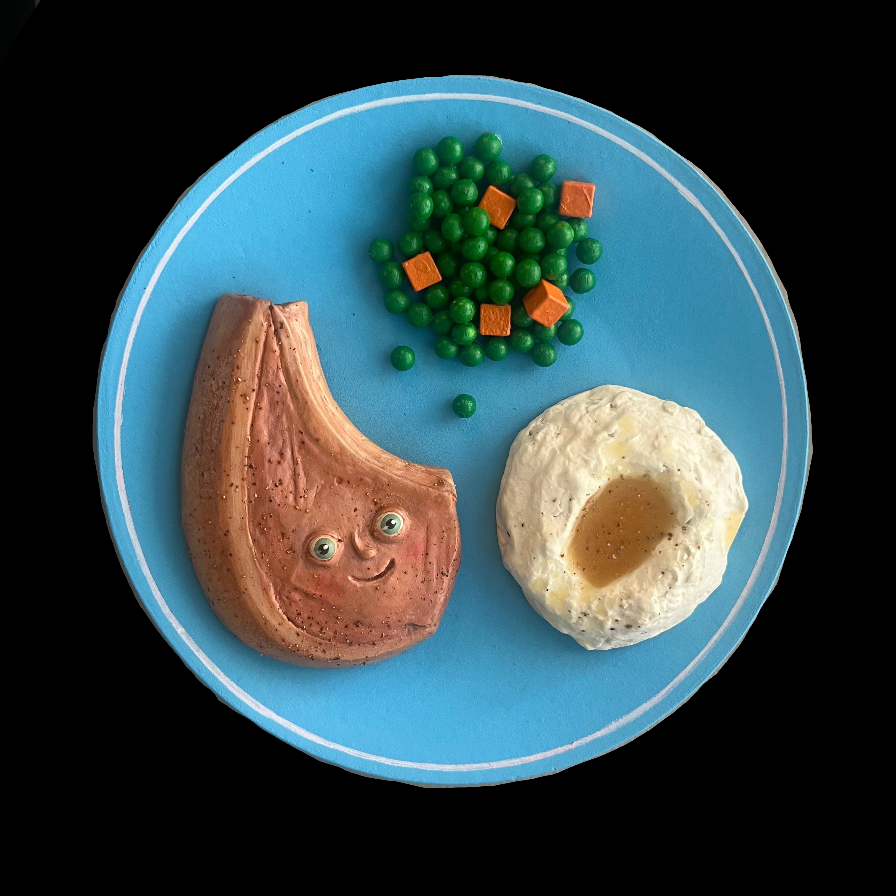 Sculpted Meat Steak with eyes next to a dollop of mashed potatoes and peas with carrot cubes on blue plate.