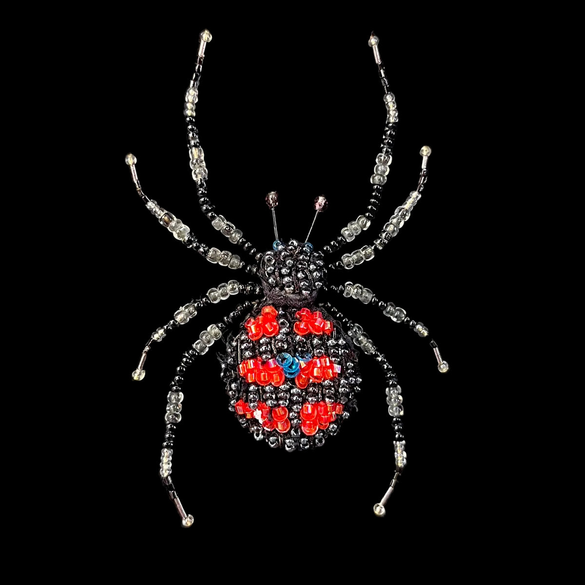 A beaded spider brooch with a black and silver body and legs, featuring red and blue rhinestone accents on its abdomen, displayed on a black velvet surface.