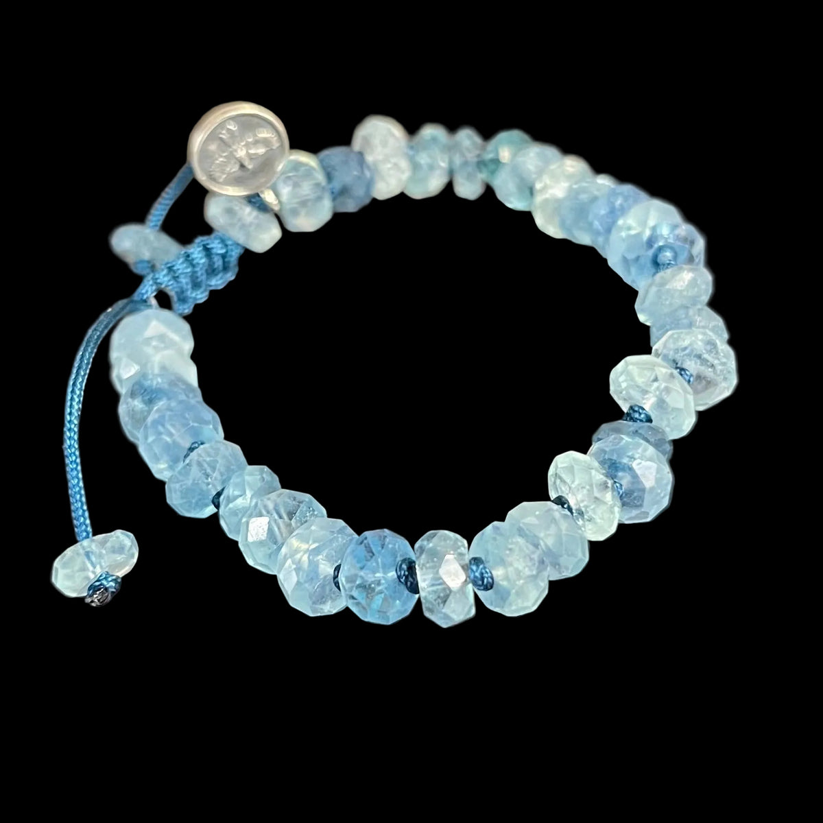 A beaded stretch bracelet made of faceted light blue beads with a blue cord and sliding knot closure, photographed on a dark reflective tabletop.