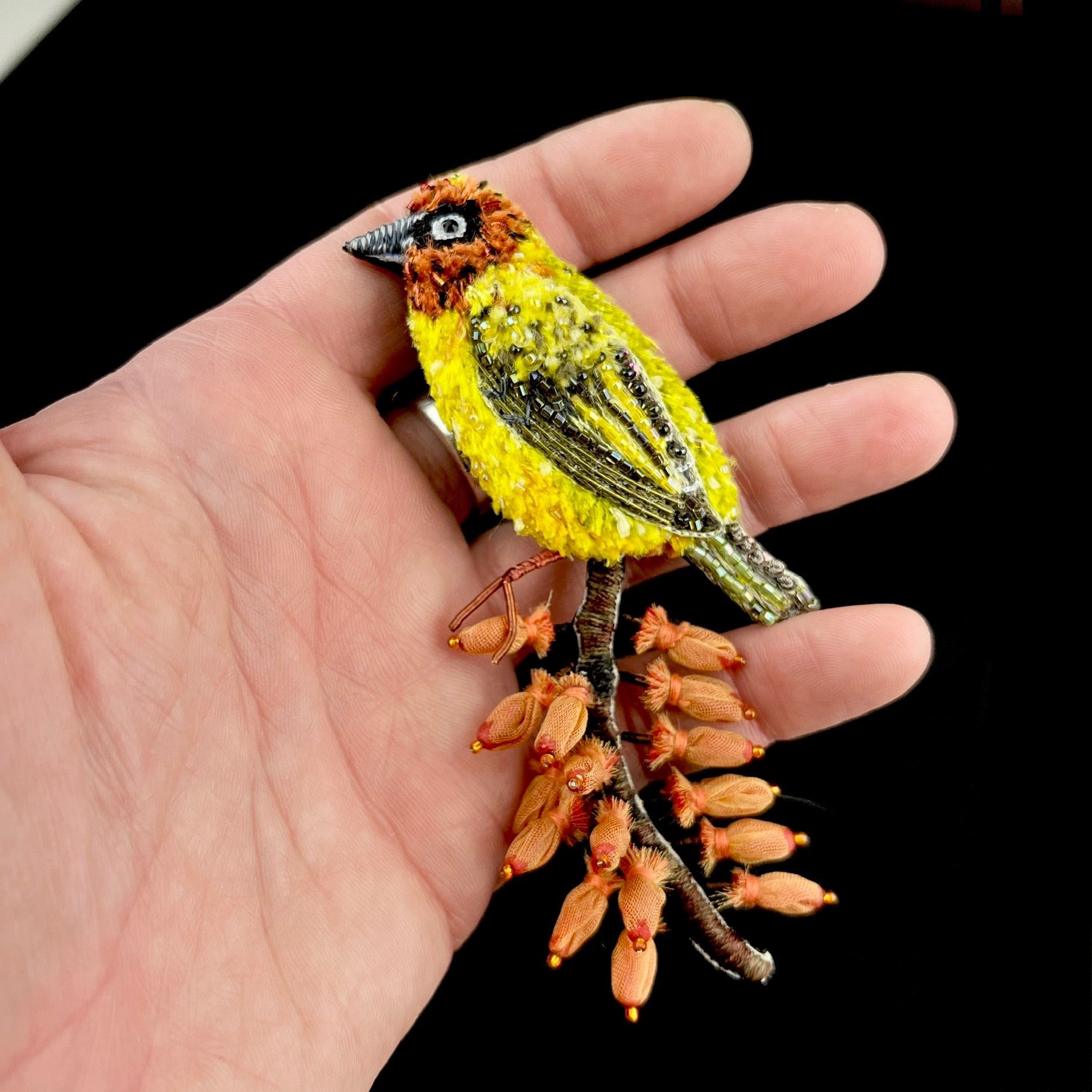 A bright yellow bird stands perched on top of a brown stick with iron colored berries which match the color of the birds head show held in a hand