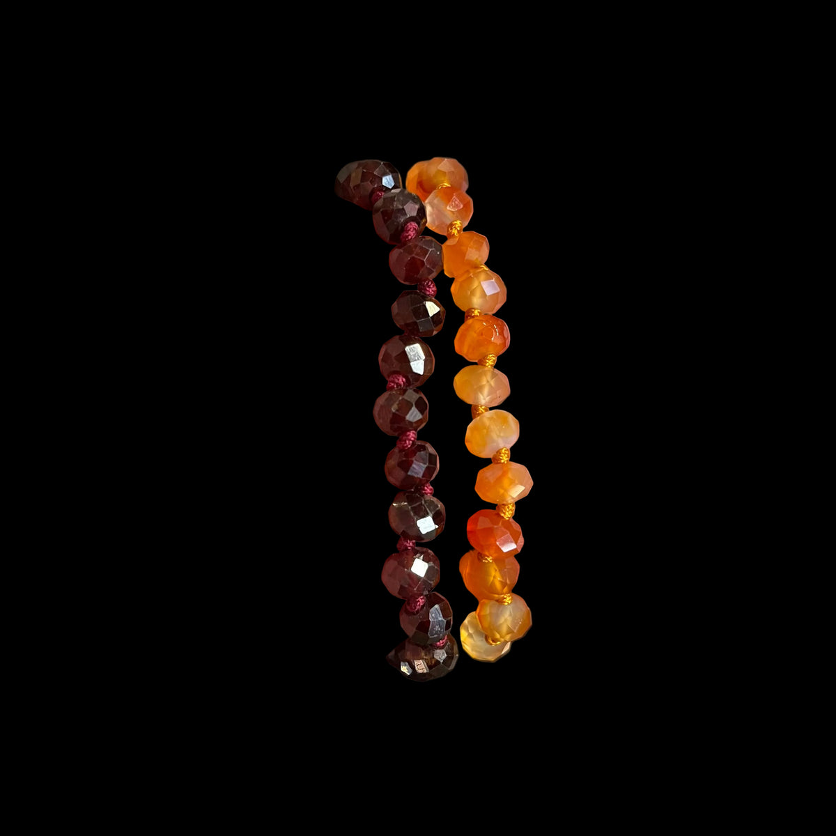Two beaded bracelets held in a hand. One bracelet features faceted dark red beads, and the other features faceted amber-orange beads.