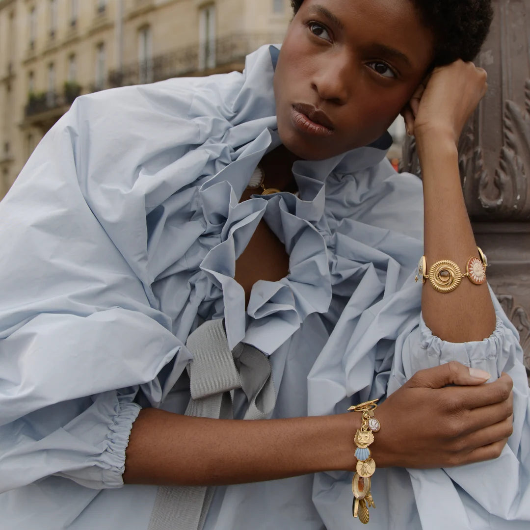 Model in blue blouse with shining gold bracelets displaying large charms set against European building