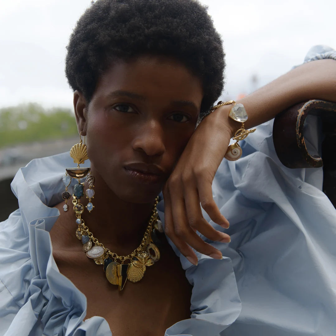 female model wearing a necklace of alternating solid gold charms with stones of different shapes and colors appearing to be found from vintage jewelry on large link gold chain with matching earrings and bracelet against blue sky and trees. 