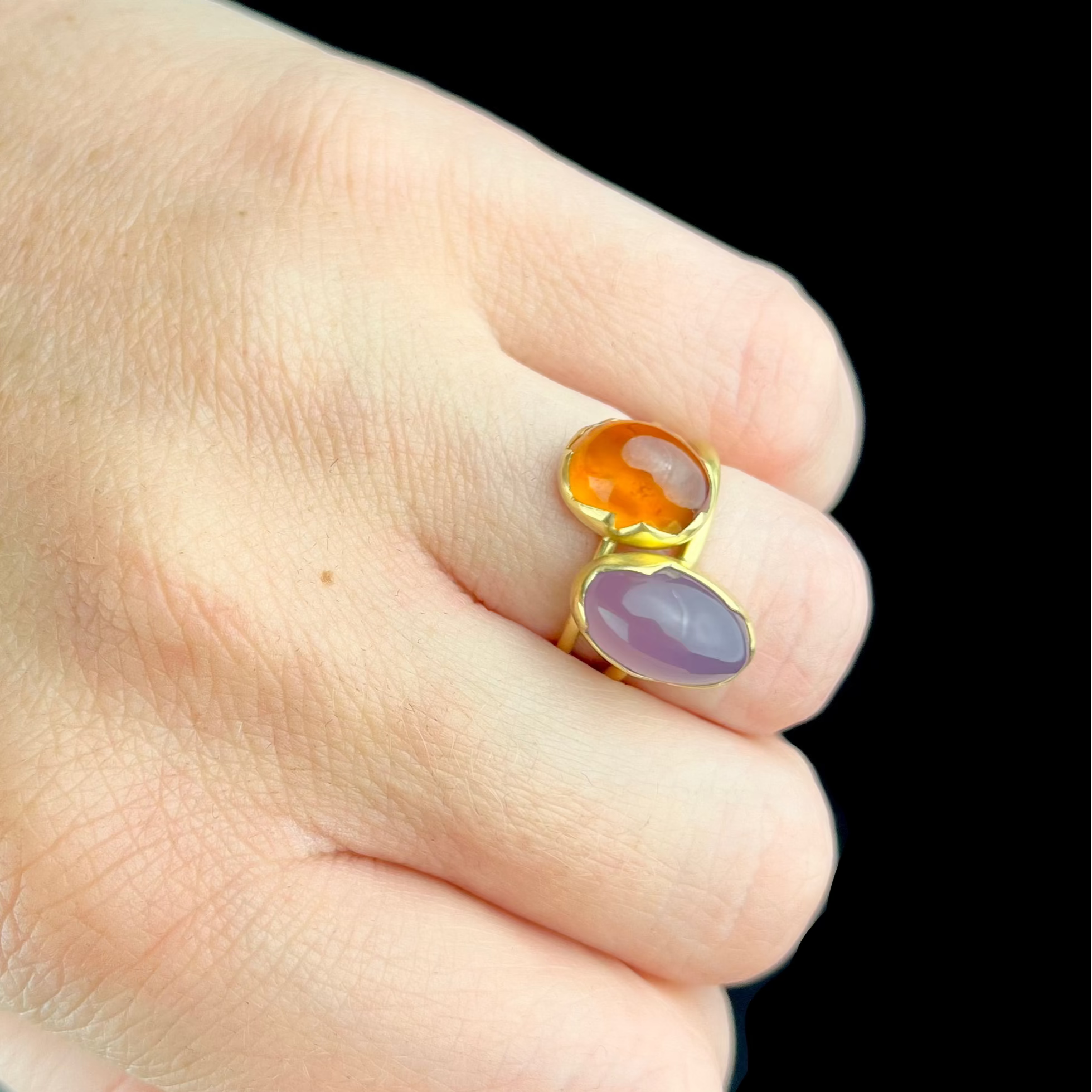 Gold ring with orange and purple stones on a finger against a dark background