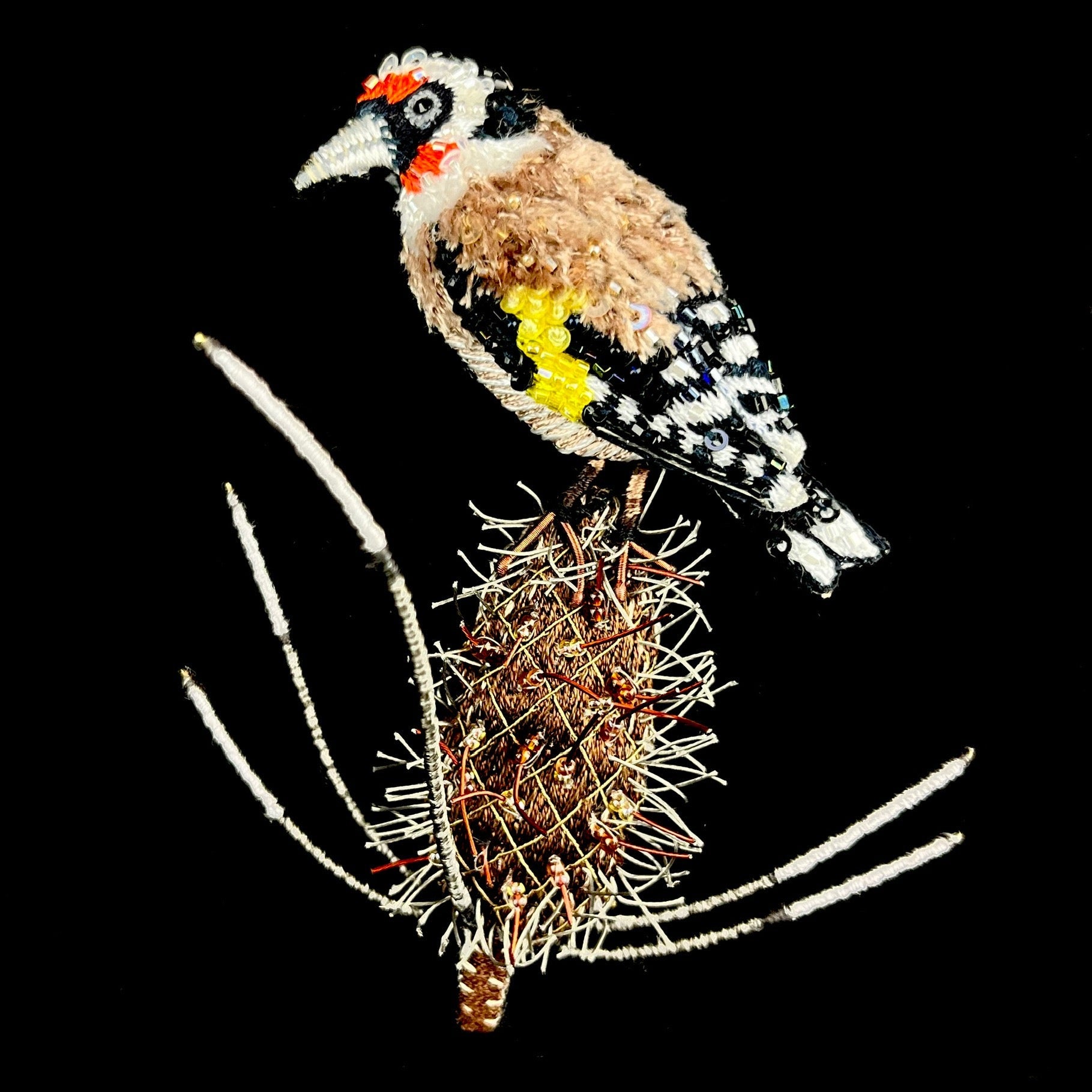 Fluffy brown,  white, black, red and yellow bird perched atop a spiked brown seed pod made of fabric and soft wire.