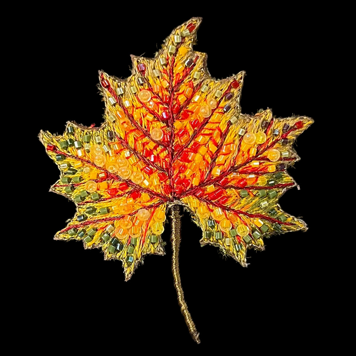 A decorative embroidered maple leaf with autumn colors, featuring yellow, orange, red, and green sequins on a black background.