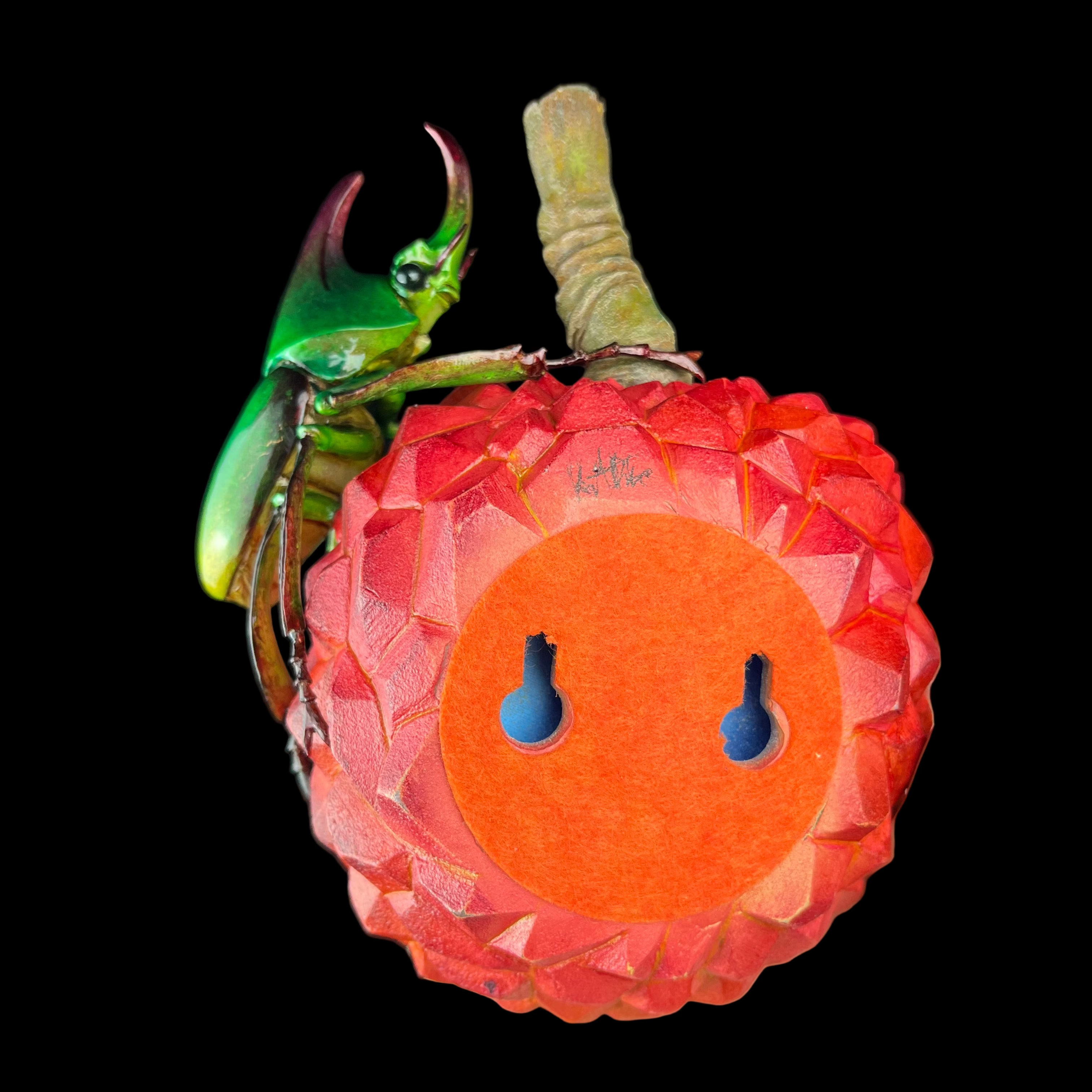 Hand holding a decorative fruit with a green insect on top against a black background