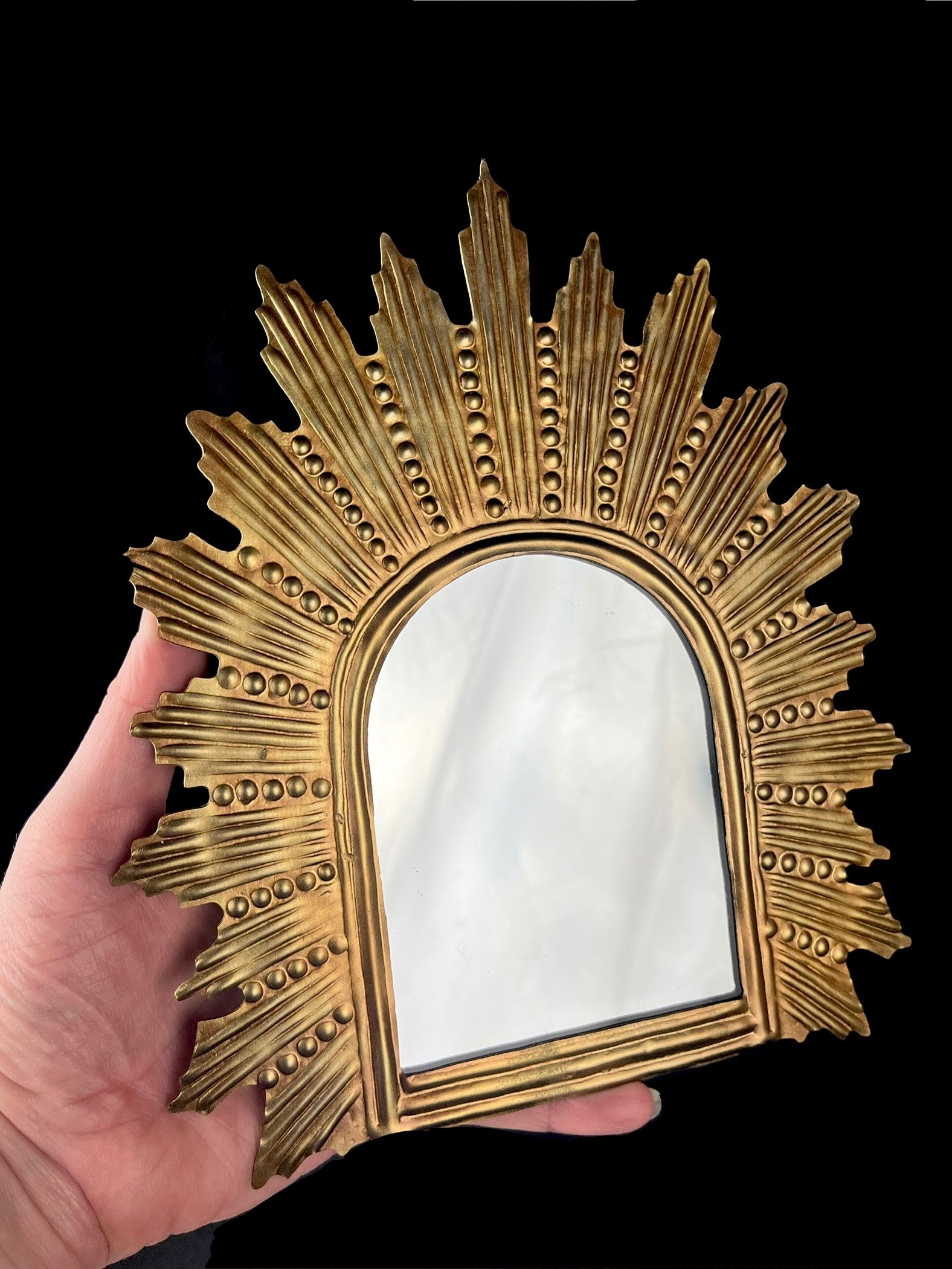 Decorative gold mirror with sunburst design held by a hand against a black background