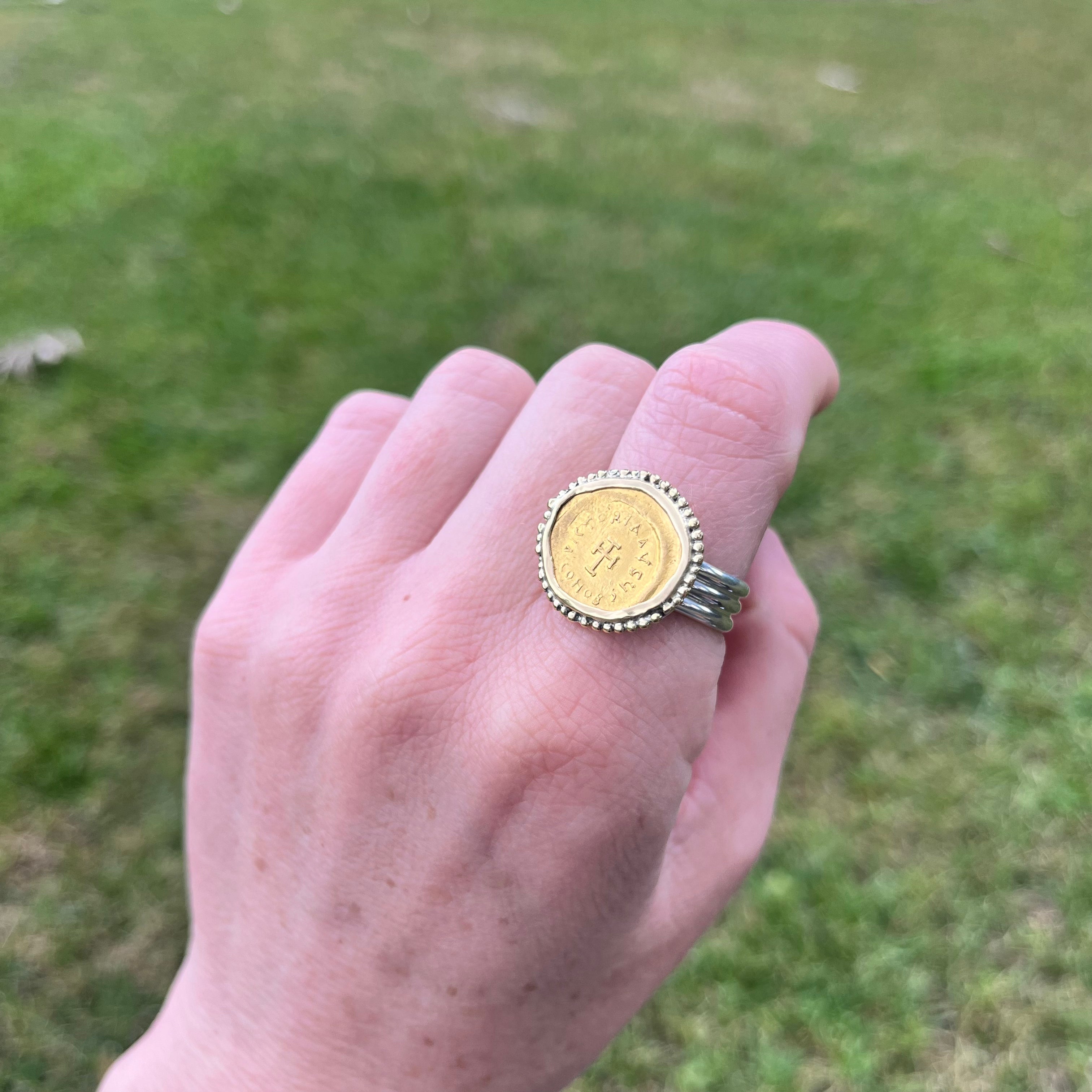 Hand wearing a gold ring with a coin design against a grassy background