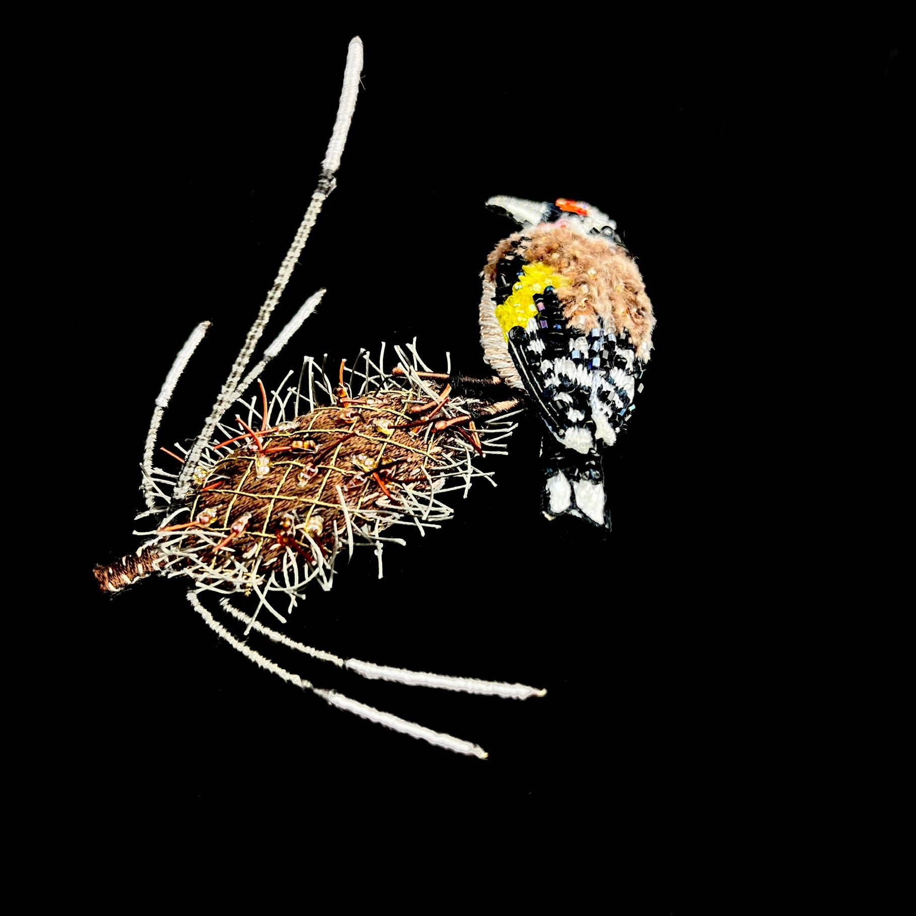 Fluffy brown,  white, black, red and yellow bird perched atop a spiked brown seed pod made of fabric and soft wire side view.
