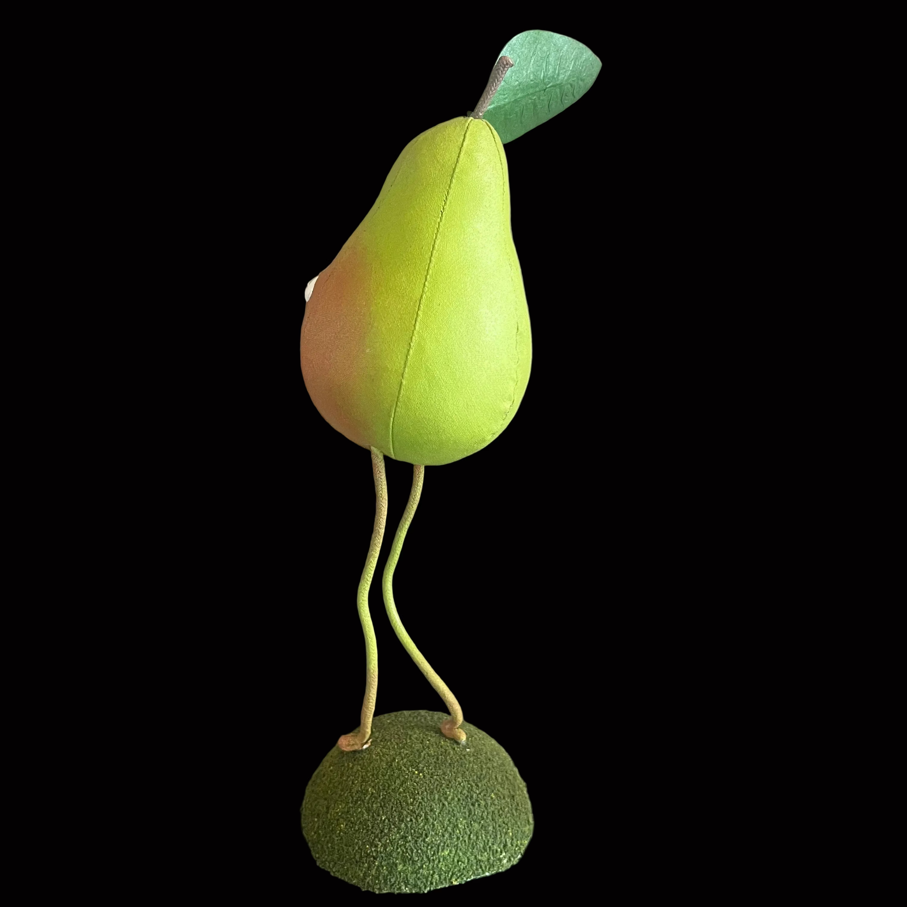 Whimsical vegetable-shaped decorative figure standing on mossy bases against black background resembling a green pear with green leaf.