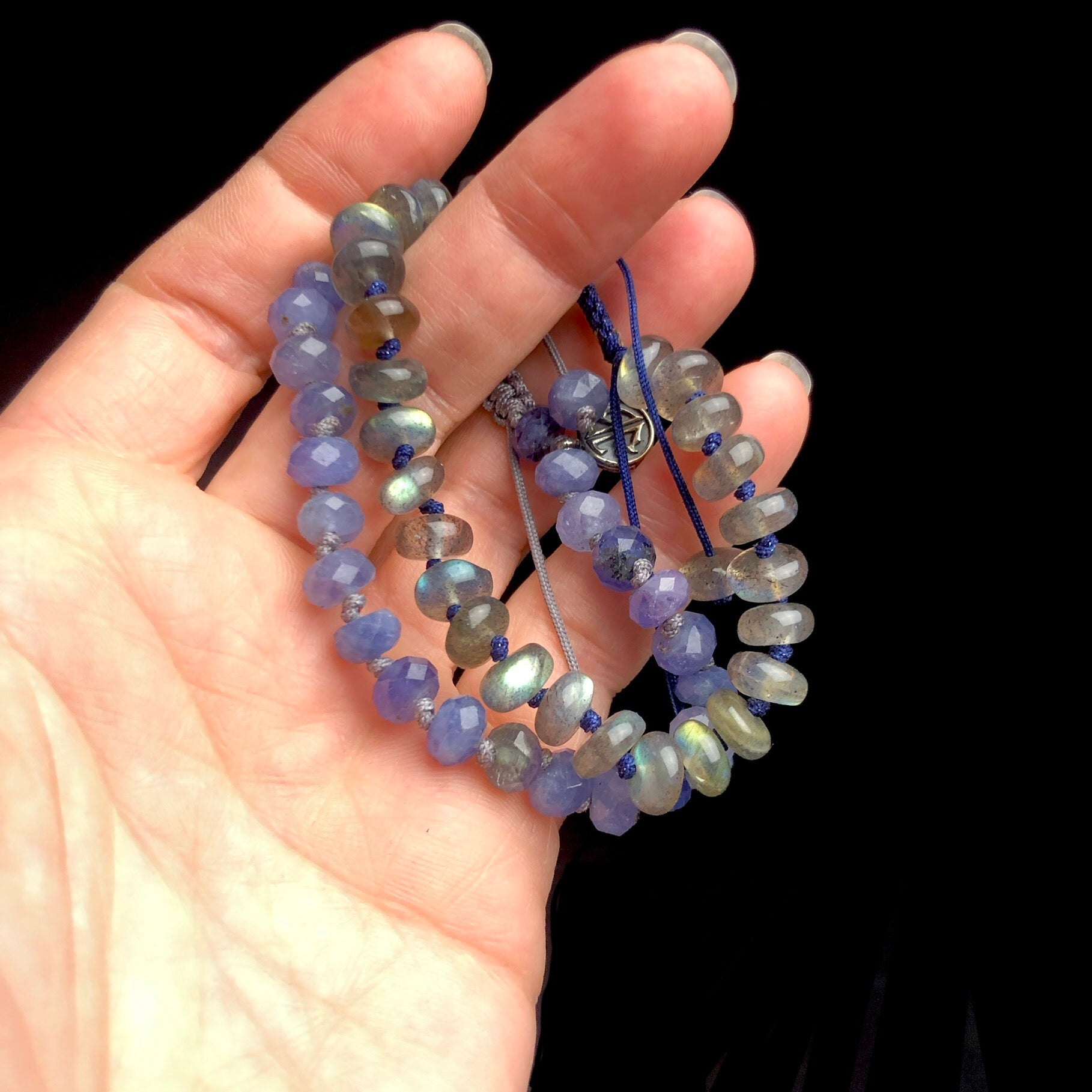 Translucent iridescent grey, blue and green colored stone bracelet with light purple knotted bracelet shown in hand 