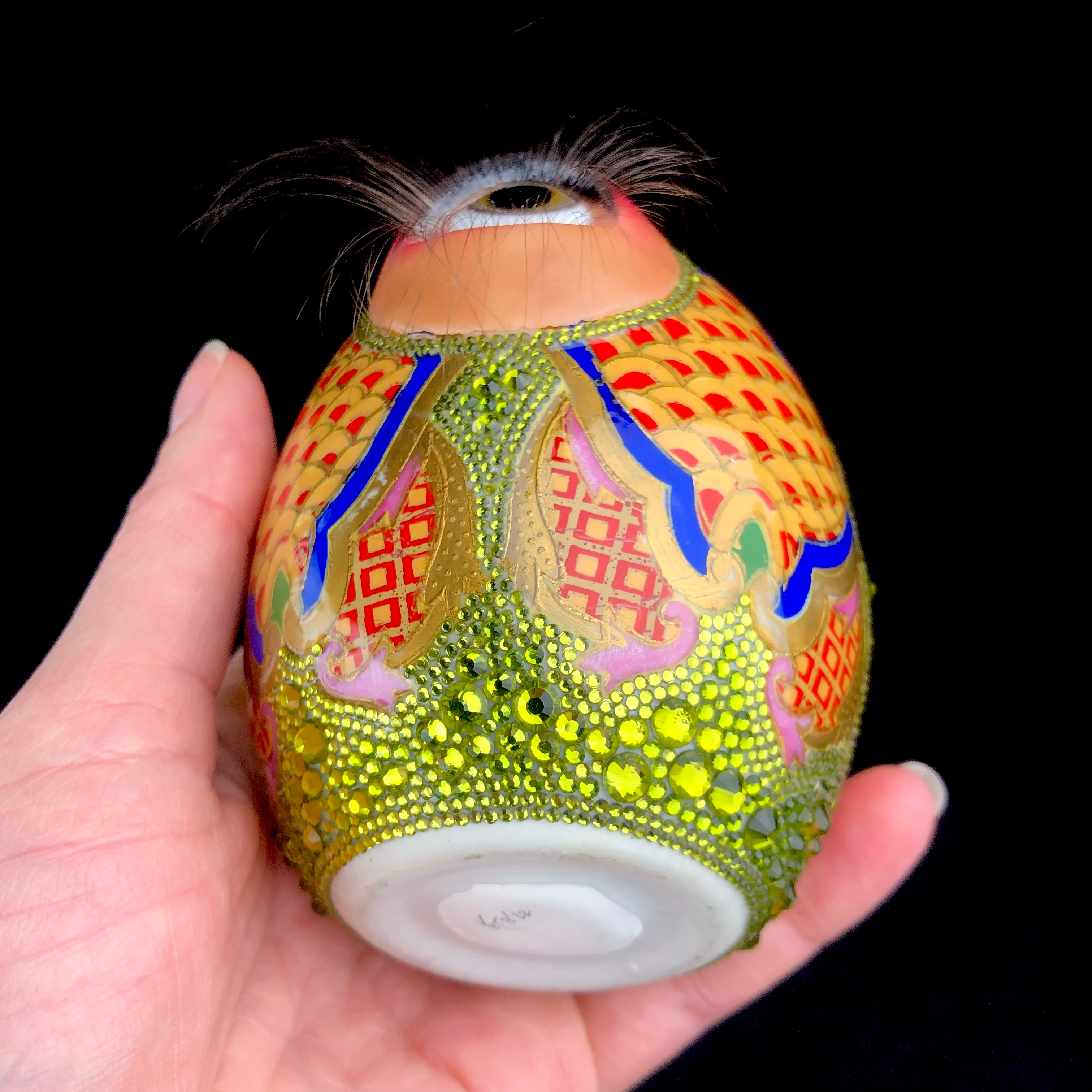 Bottom view of shiny glazed yellow, red, pink, blue and green egg with green crystals and glass human eye looking up towards long brown lashes
