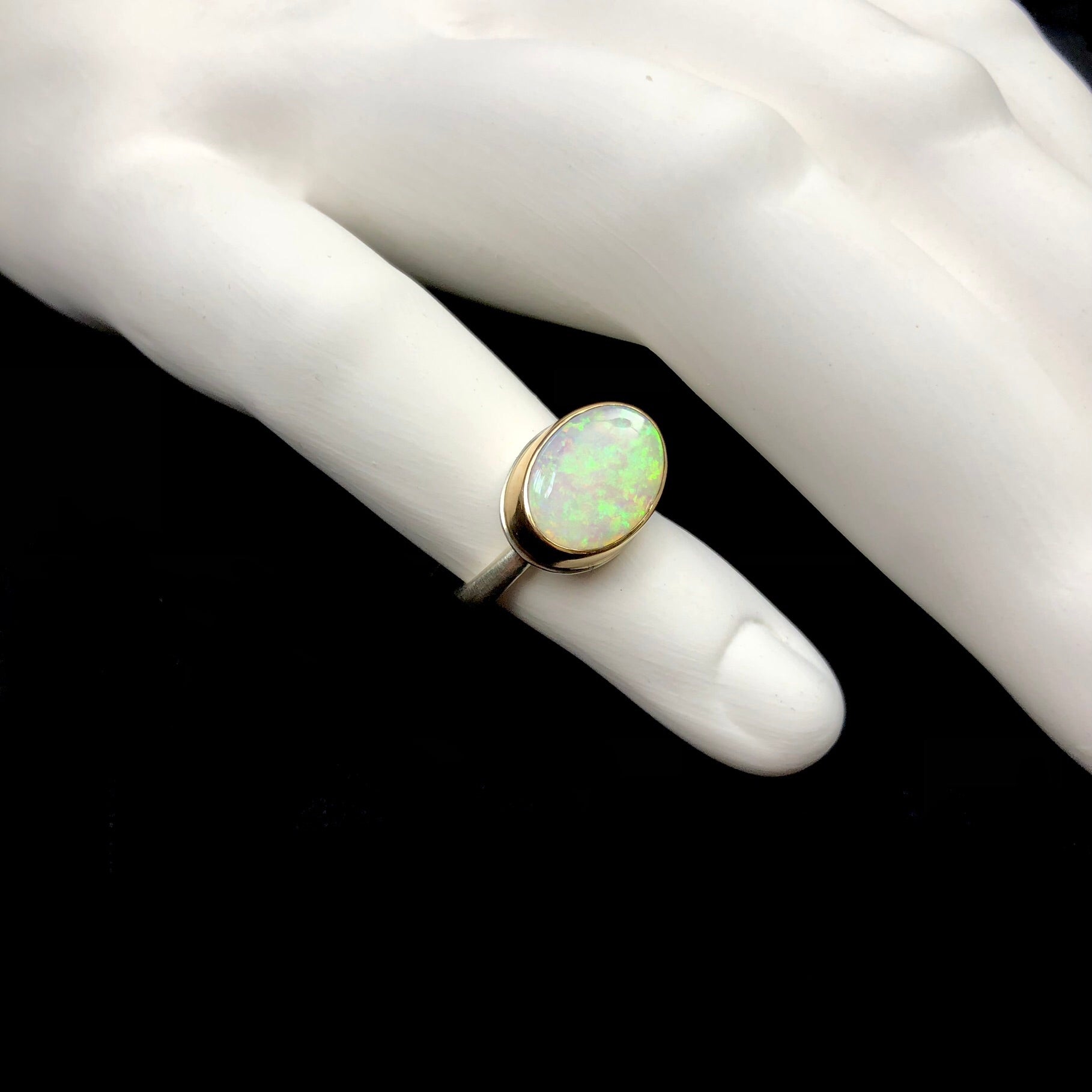 Side view of Mintabe opal stone ring with iridescent green accents, gold setting and silver band shown on shite ceramic finger