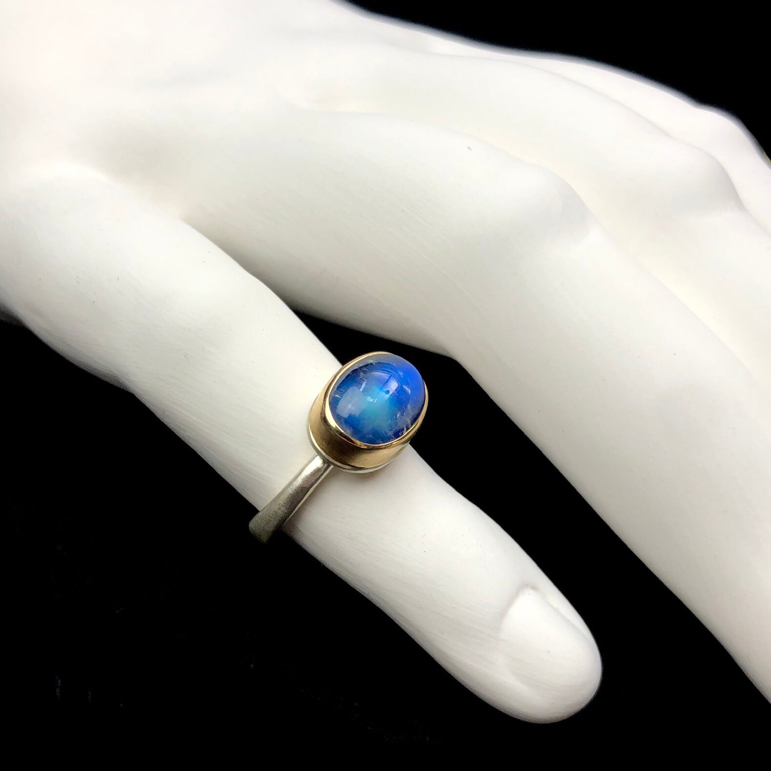 Profile view of smoothly polished blue stone with gold setting and white band shown on white ceramic finger