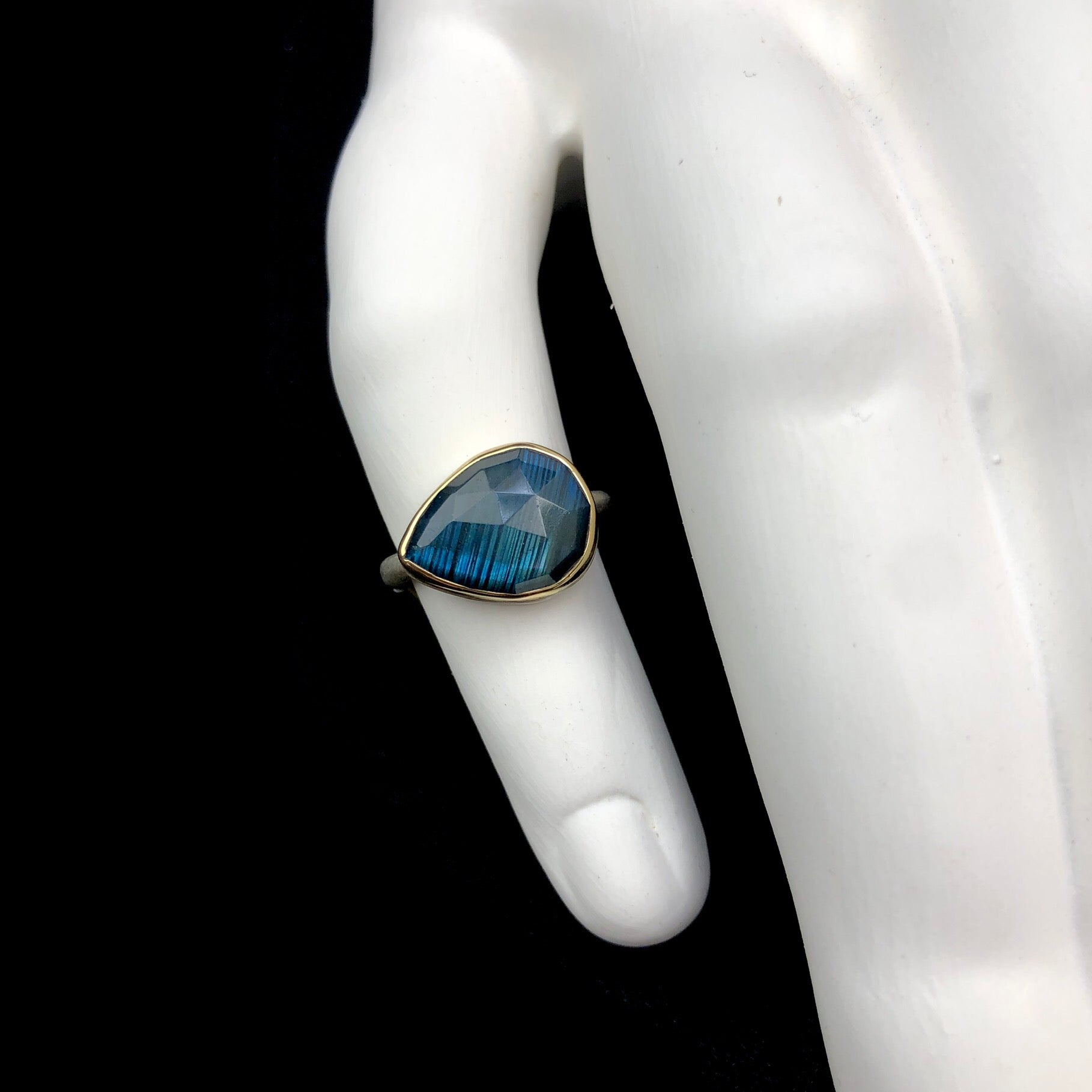 Teardrop shaped blue/green iridescent stone ring with gold setting shown on white ceramic finger