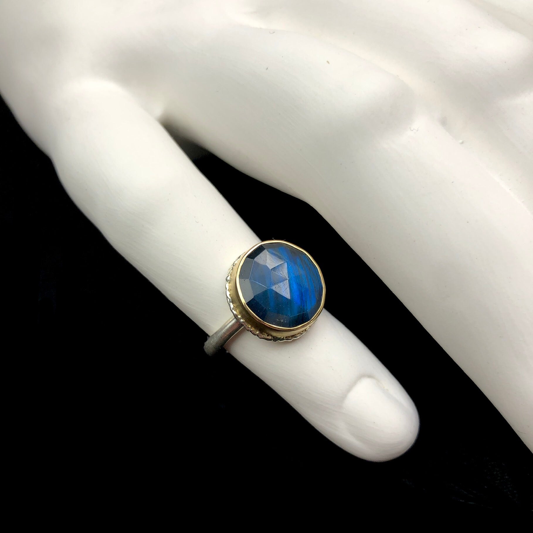 Round iridescent blue/green stone ring with gold setting shown on white ceramic finger