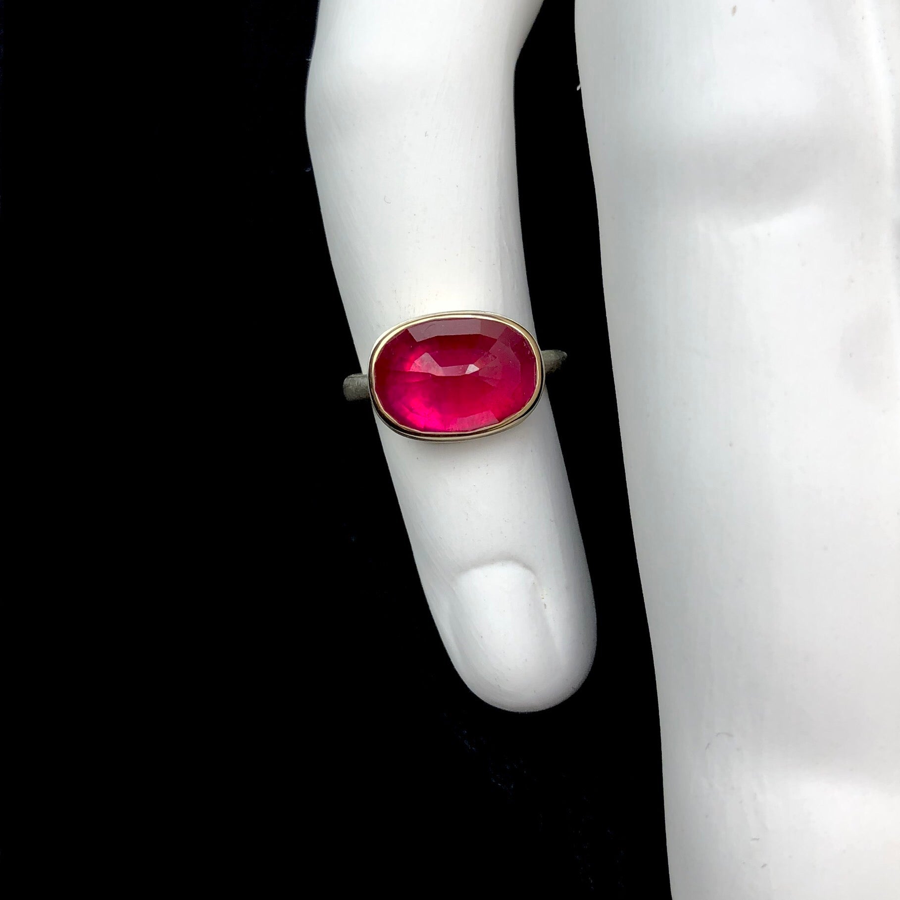 Top view of oval shaped pink/red colored ruby ring shown on white ceramic finger