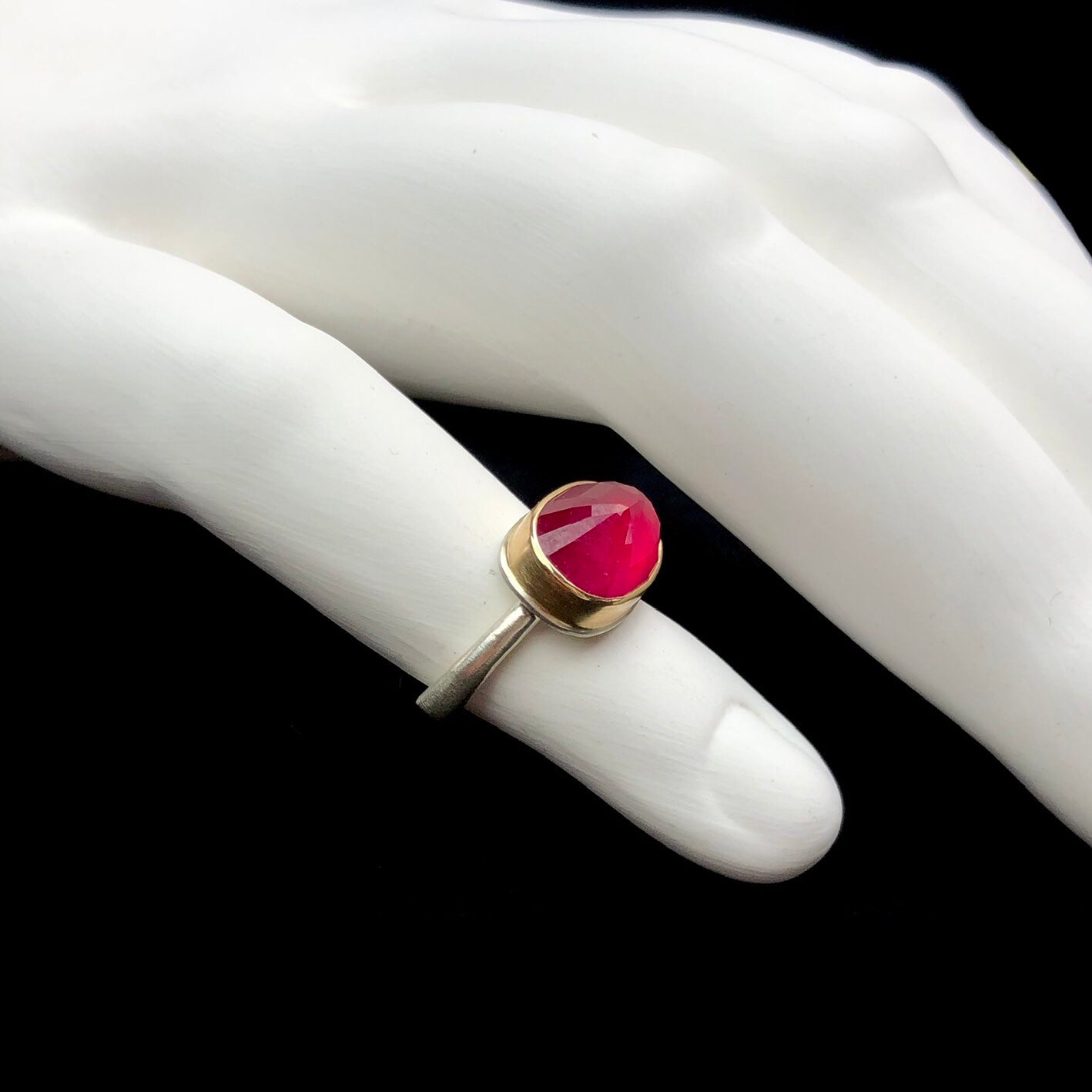 Side profile view of faceted, raised pink/red colored ruby stone ring shown on white ceramic finger