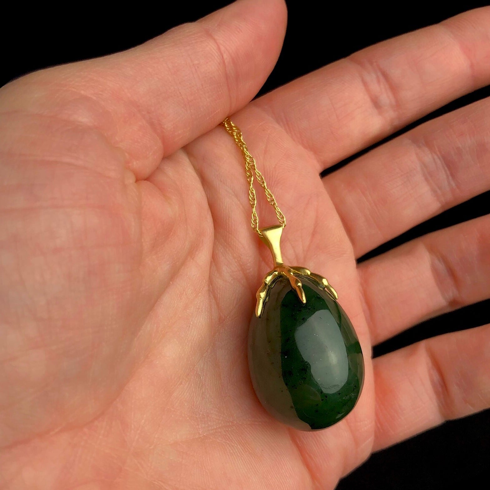 Green egg shaped stone held by a gold bird claw on gold chain shown in hand