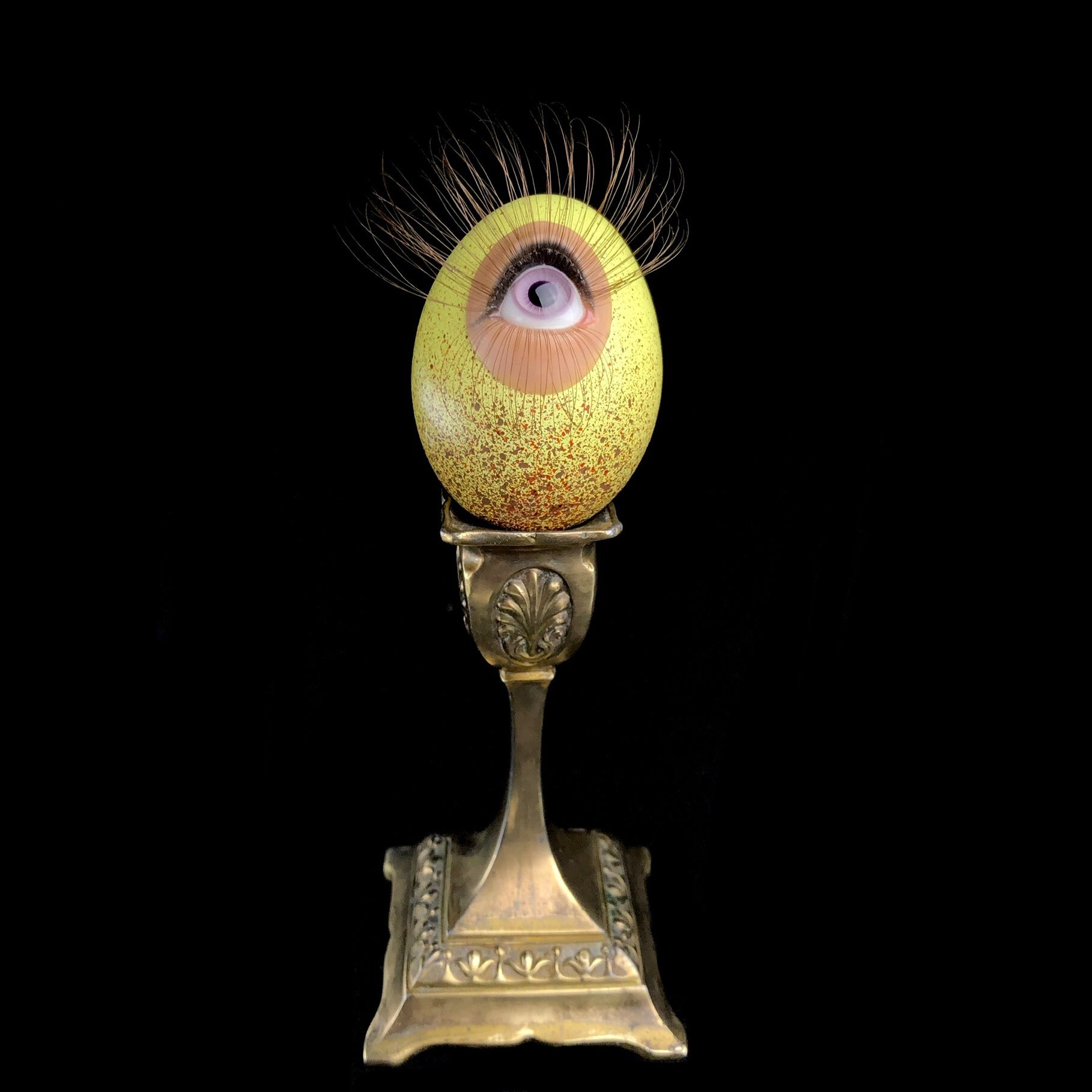 Front view of yellow egg with purple eye and brown eyelashes sitting on brass stand