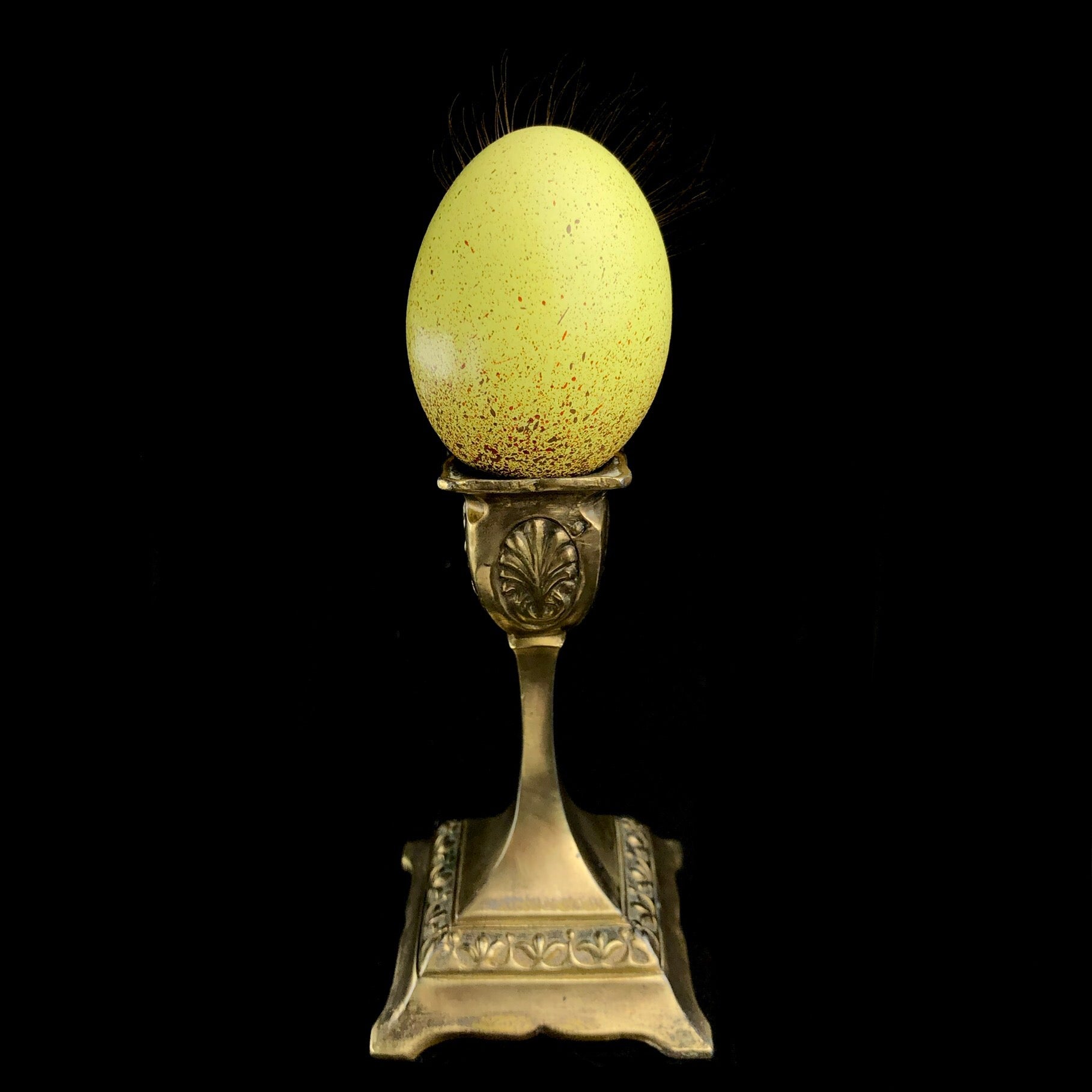 The back view of Verona the Chicken Egg, yellow with brown spots sitting on brass stand