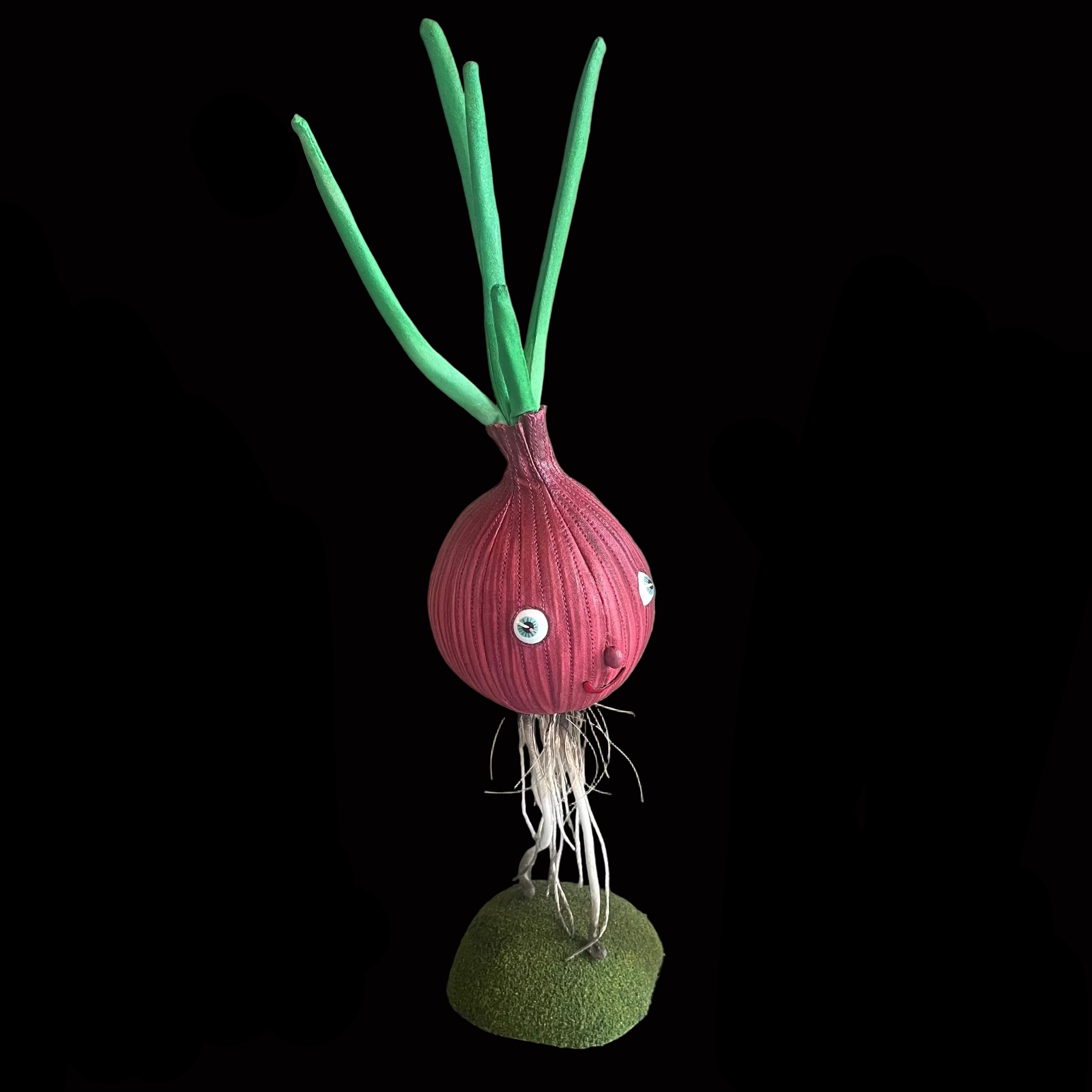 Whimsical vegetable-shaped decorative figure standing on mossy bases against black background resembling a red onion with green leaves and a smiling face.