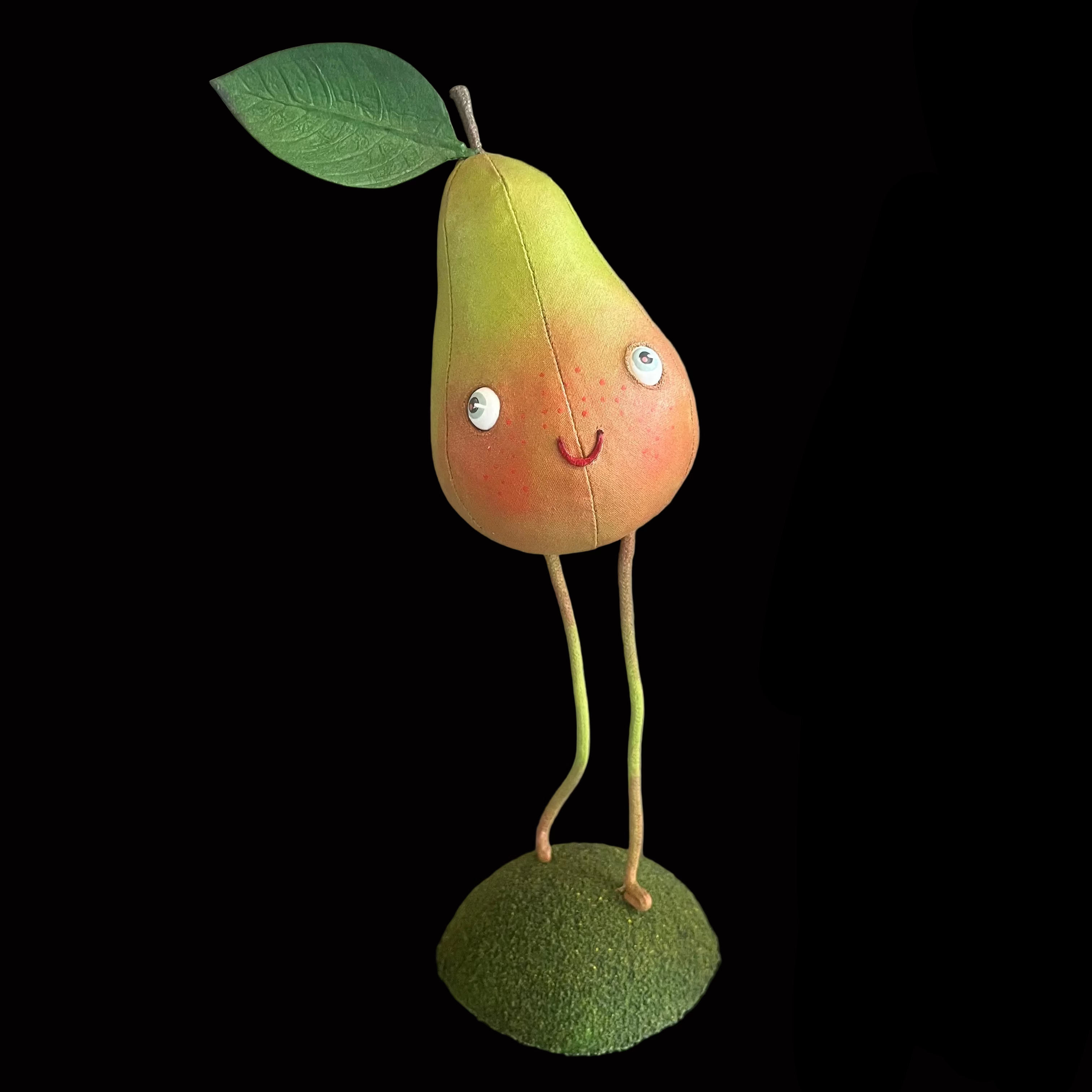 A handmade plush pear character toy with a green leaf on its head, standing on a mossy base. The pear has a yellow-to-orange gradient body with embroidered eyes, a smile, and long thin legs.
