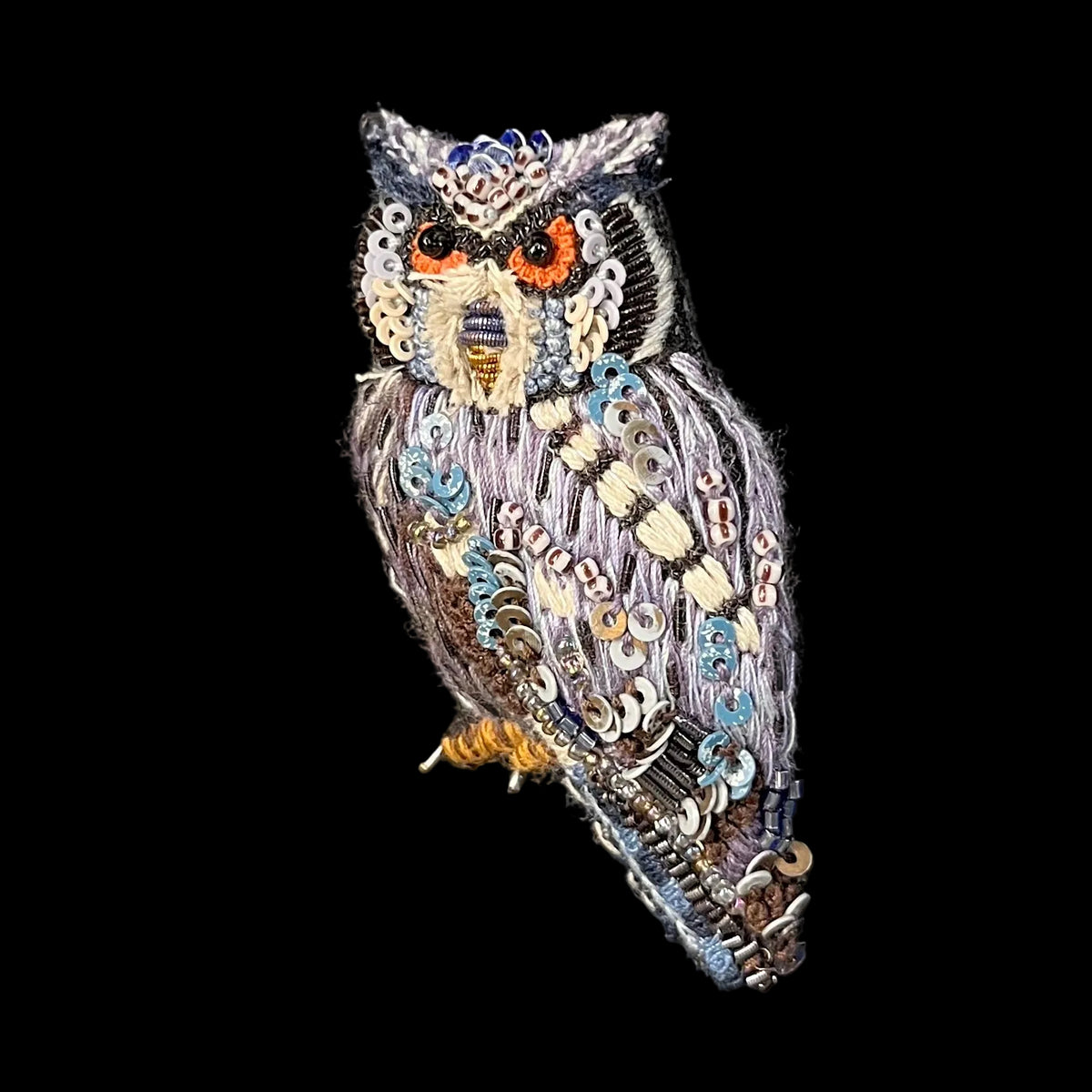 A highly detailed, handcrafted owl brooch or patch featuring intricate beadwork, sequins, and thread embroidery in shades of gray, blue, white, orange, and yellow, set against a solid black background.
