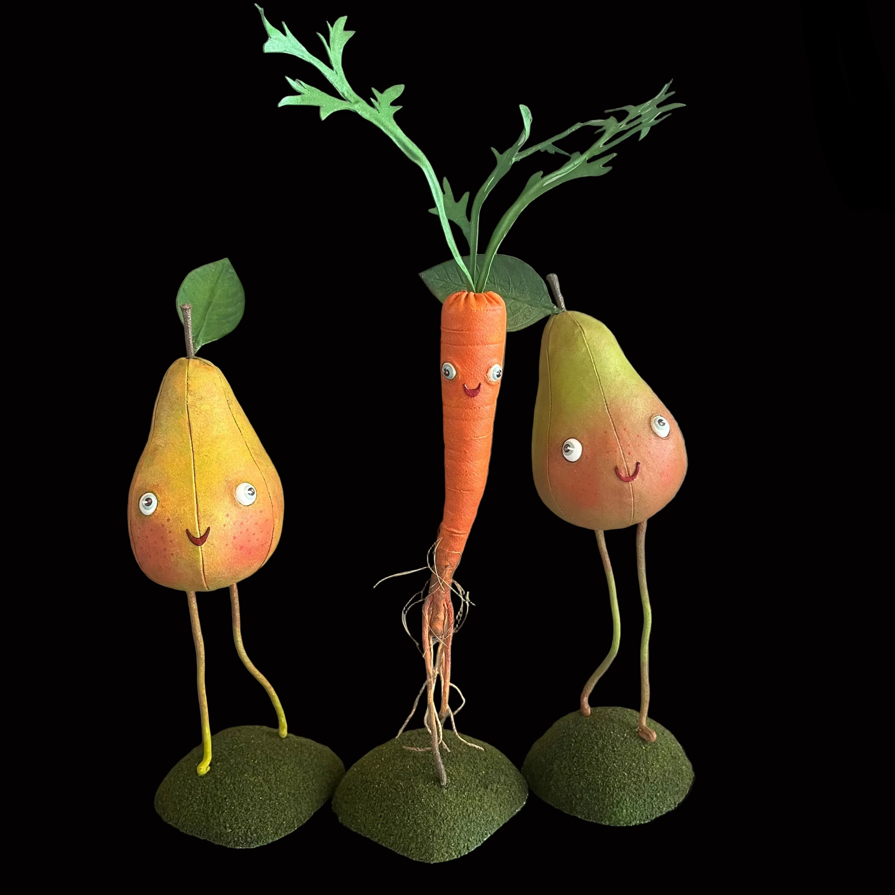 Whimsical vegetable-shaped decorative figures standing on mossy bases against black background resembling two green pears and an orange carrot with green leaves and smiling faces.