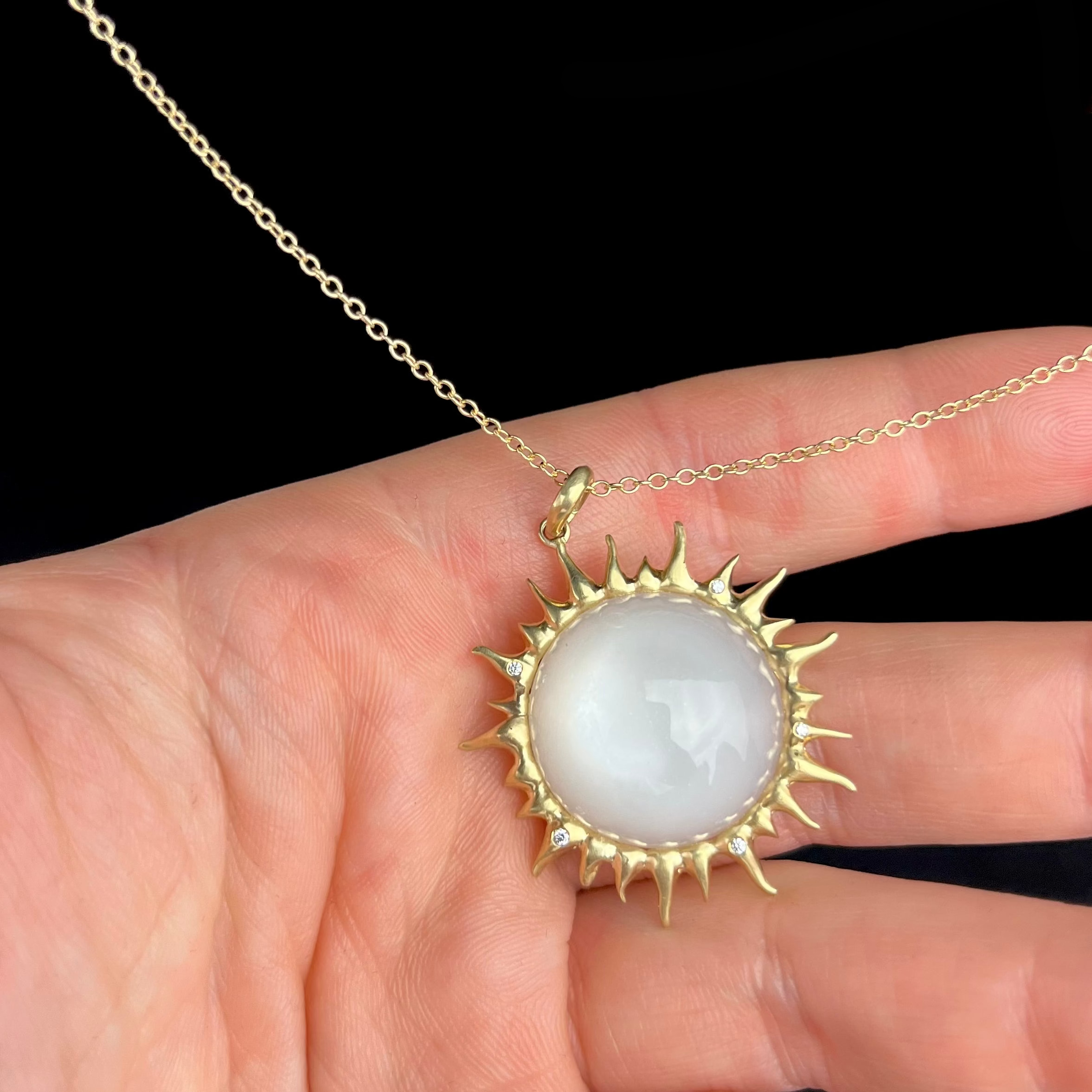 Gold necklace with a sun-shaped pendant featuring a white stone held by a hand against a black background