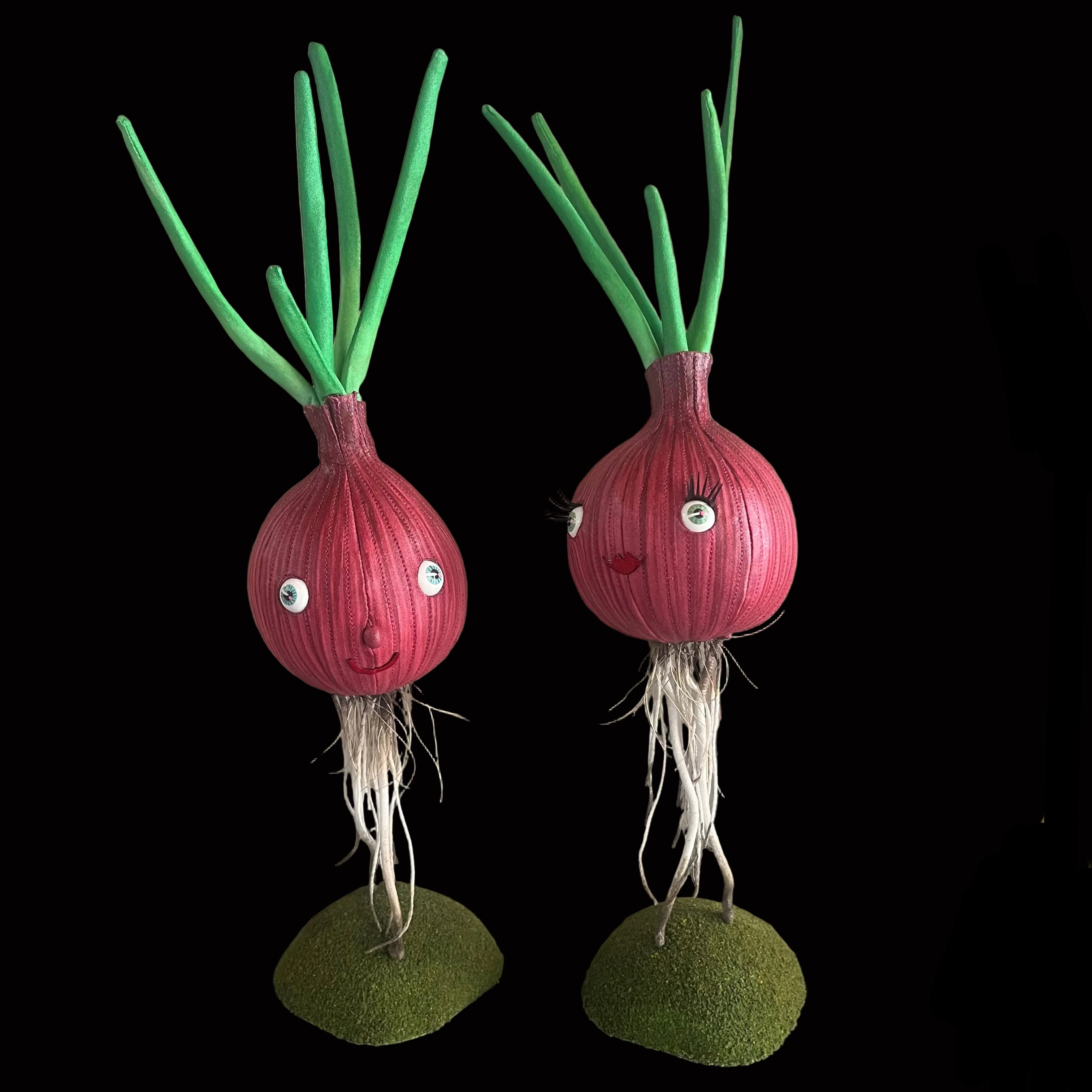 Two handcrafted fabric vegetable sculptures resembling red onions with green sprouts and white roots, displayed on small green bases against a black background.