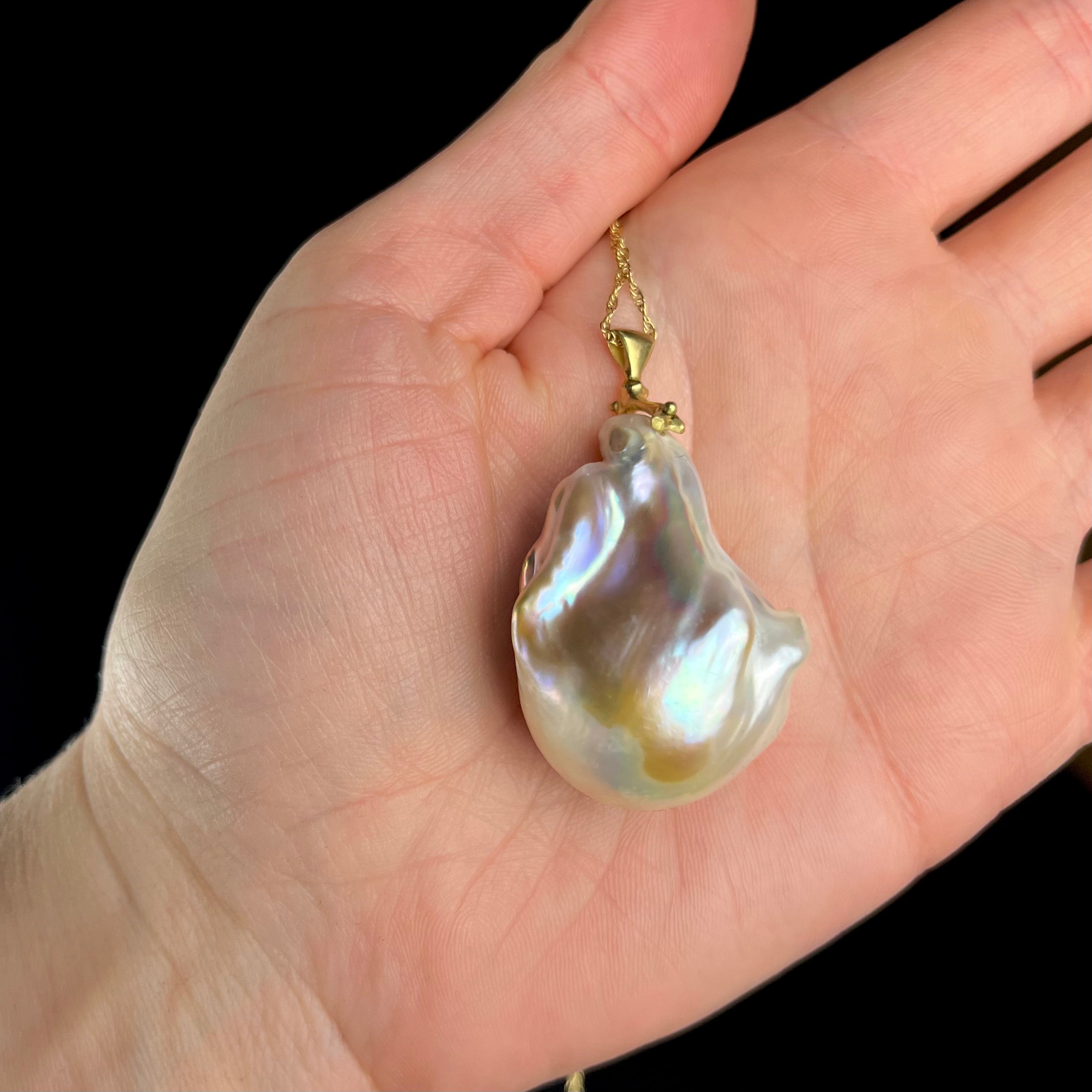 Baroque pearl pendant on a gold chain held in a hand against a black background