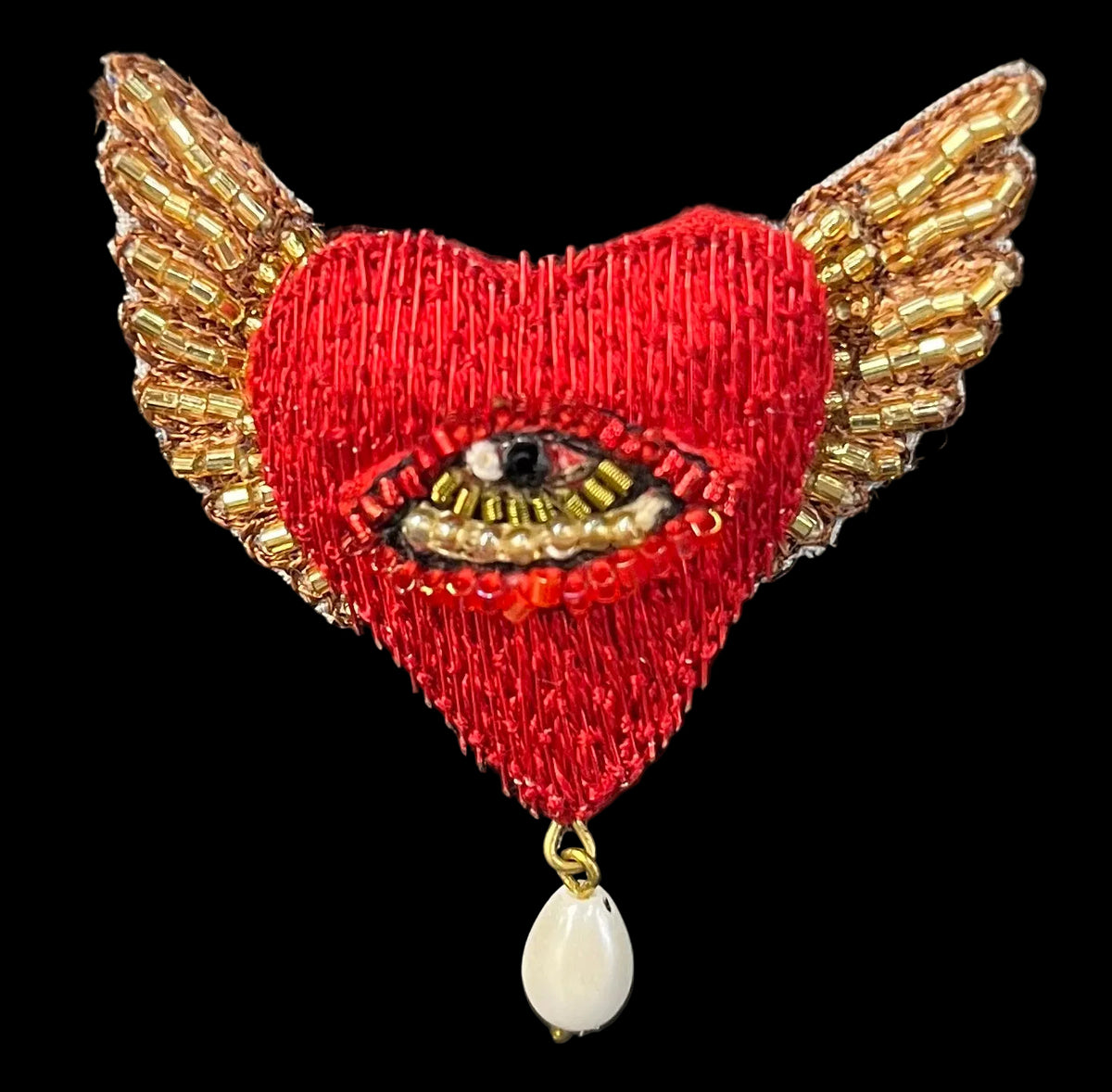 A red embroidered heart brooch with gold beaded wings, an all-seeing eye motif in the center, and a dangling white teardrop bead, shown on a black background.